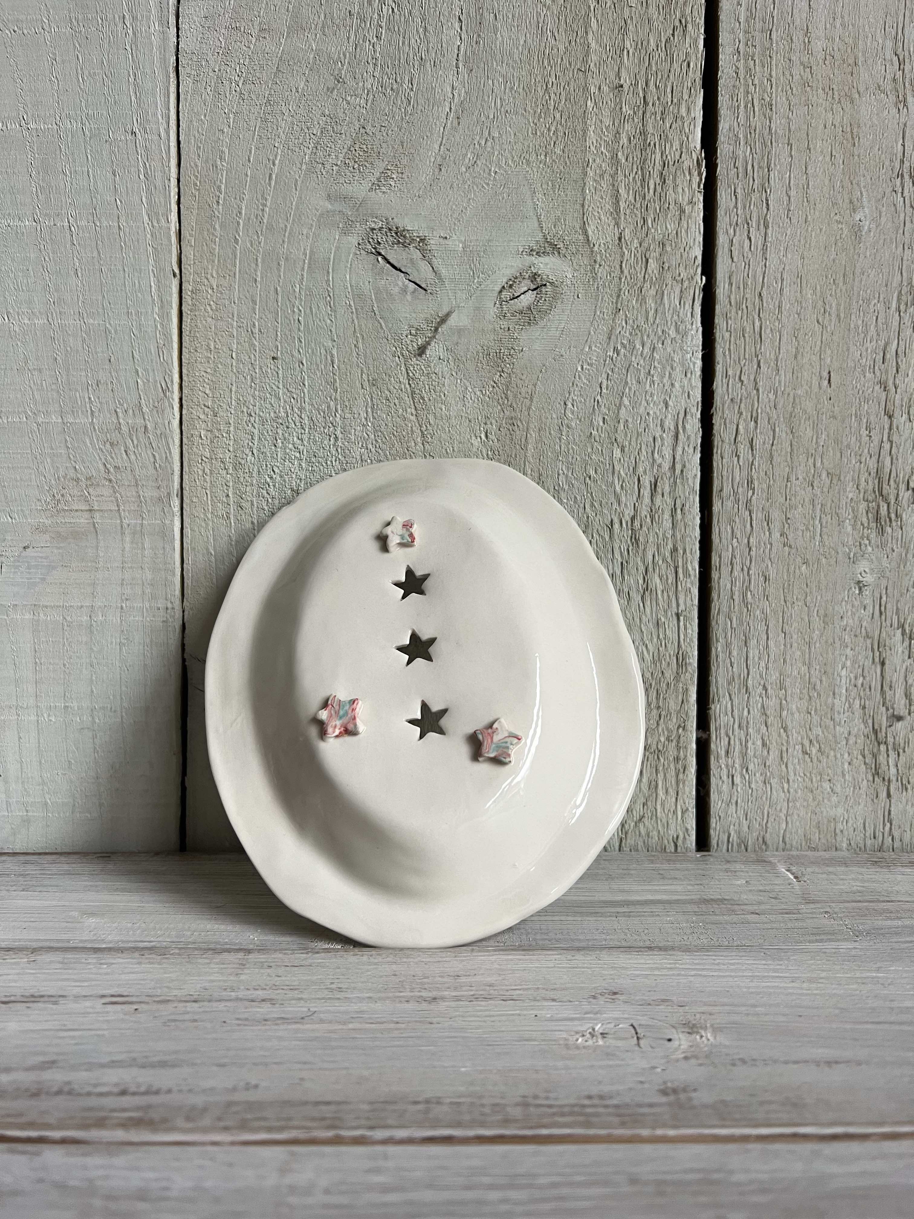White handmade ceramic trinket dish with pink leaf detailing and decorative star-shaped cut-outs standing on a rustic wooden shelf.