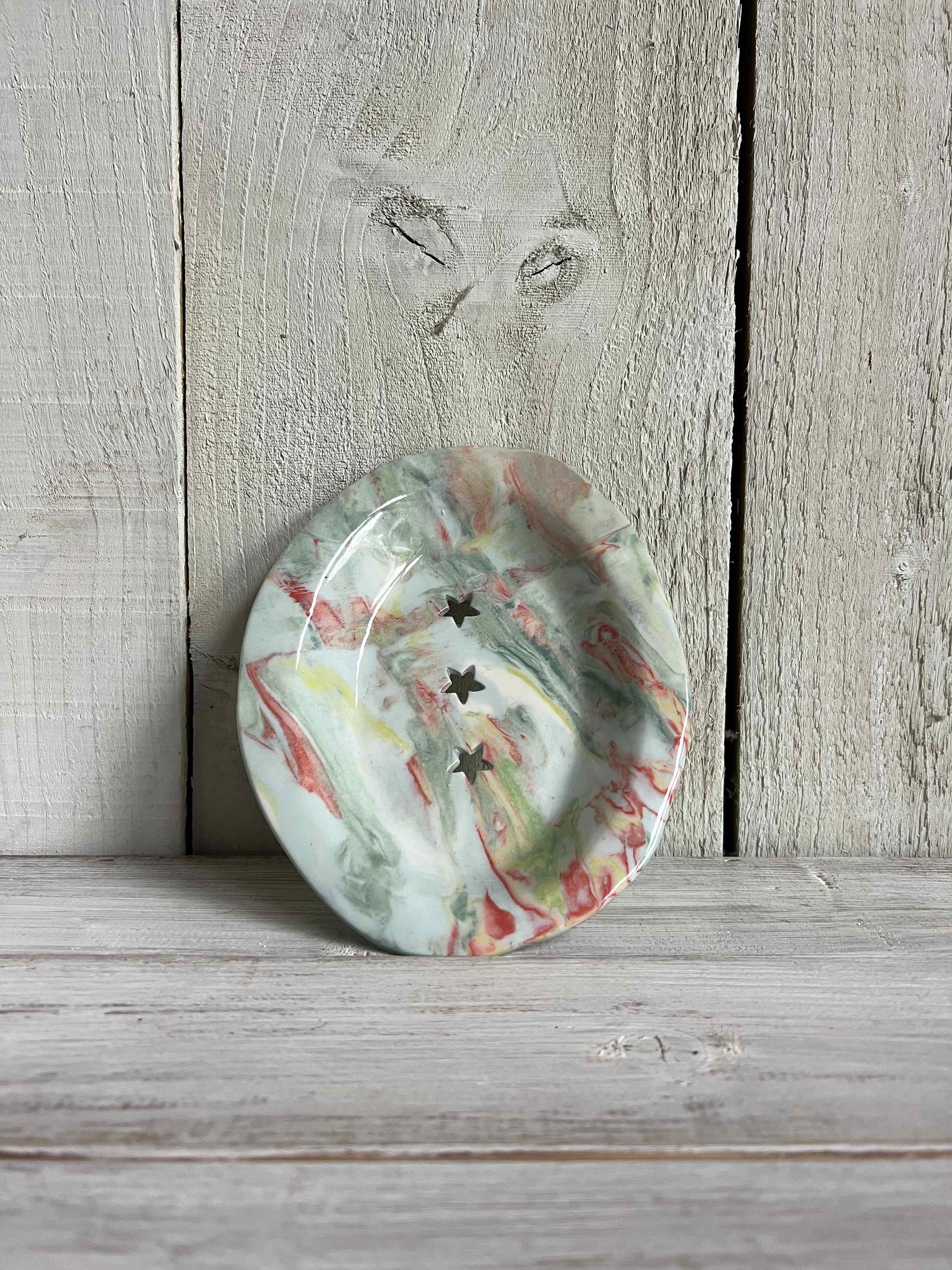 Handmade oval ceramic trinket dish with a pastel marbled glaze and three star-shaped cut-outs displayed on a rustic wooden shelf.