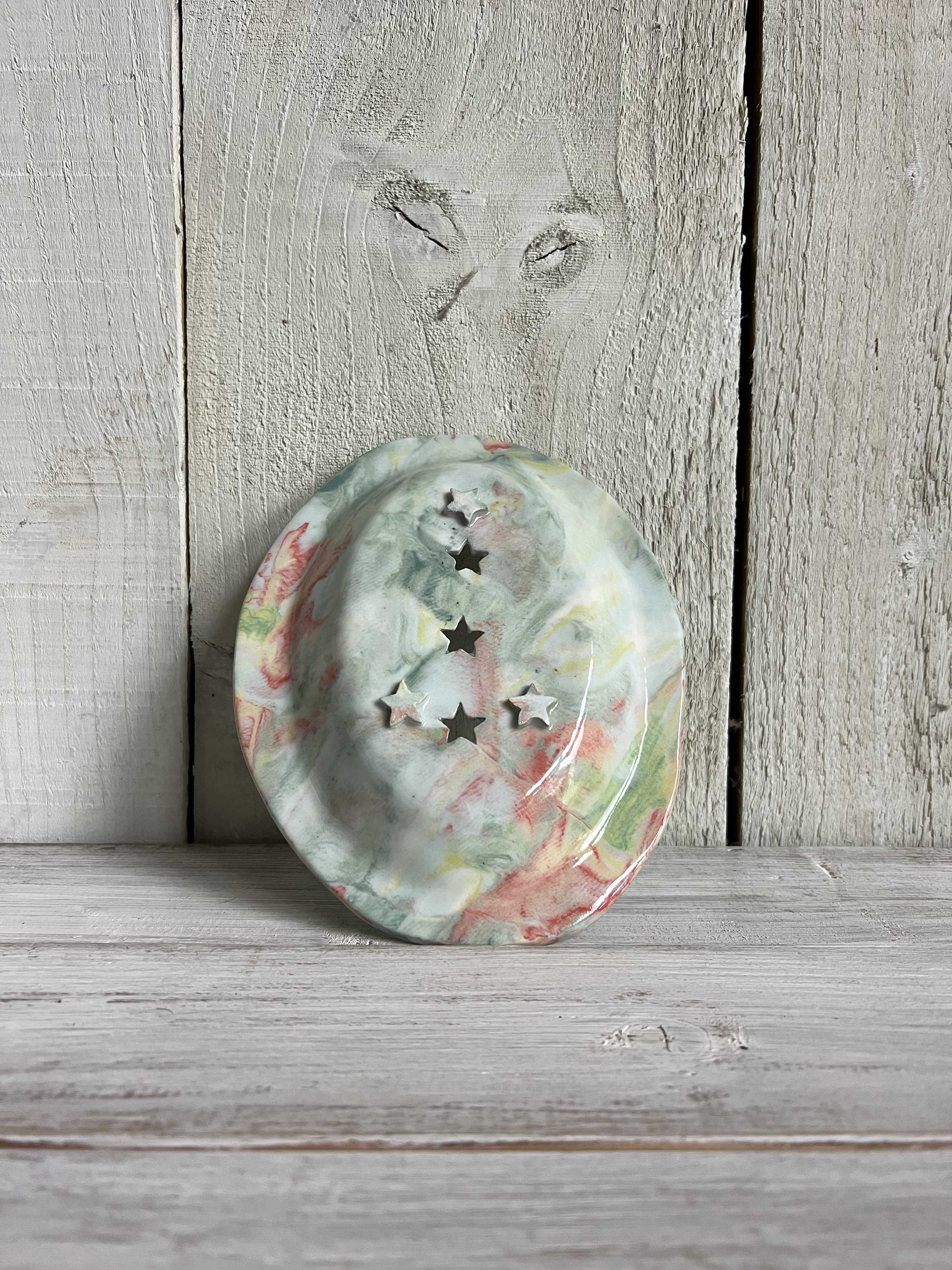 Pastel marbled oval ceramic trinket dish with three star-shaped cut-outs standing on a rustic wooden shelf.