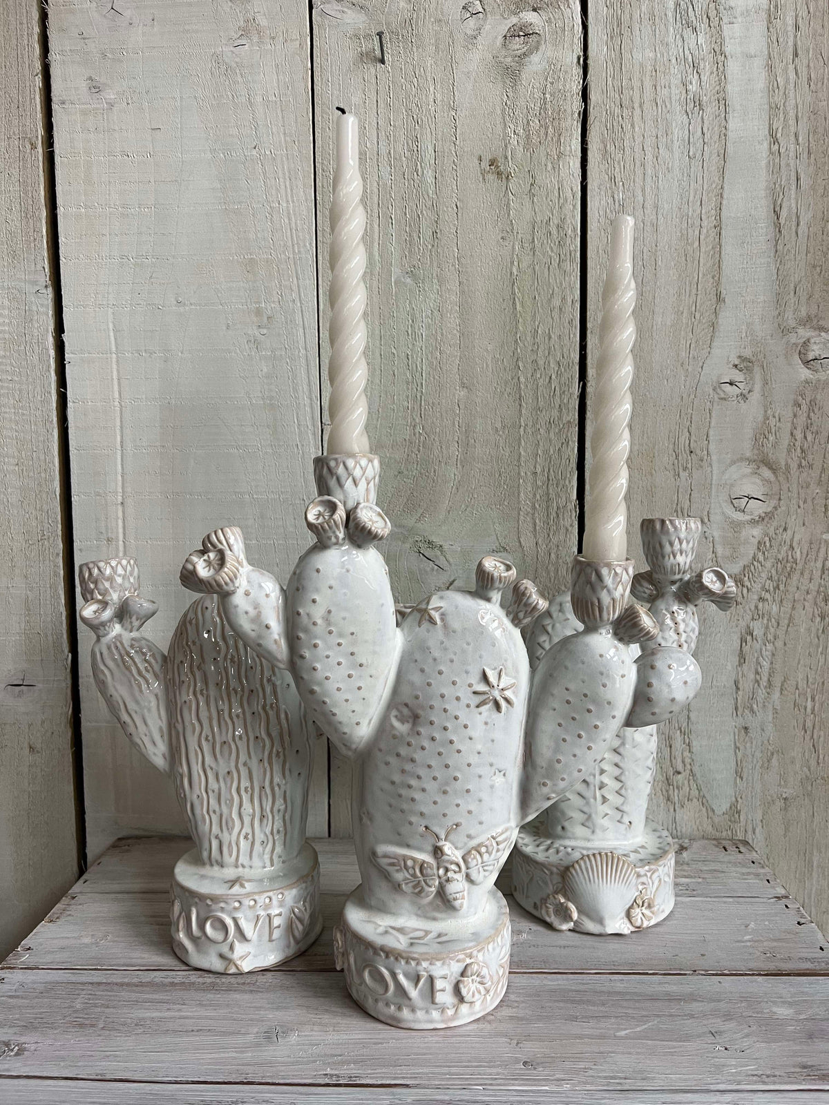 Collection of handmade white ceramic cactus candle holders displayed with taper candles on a rustic wooden shelf.