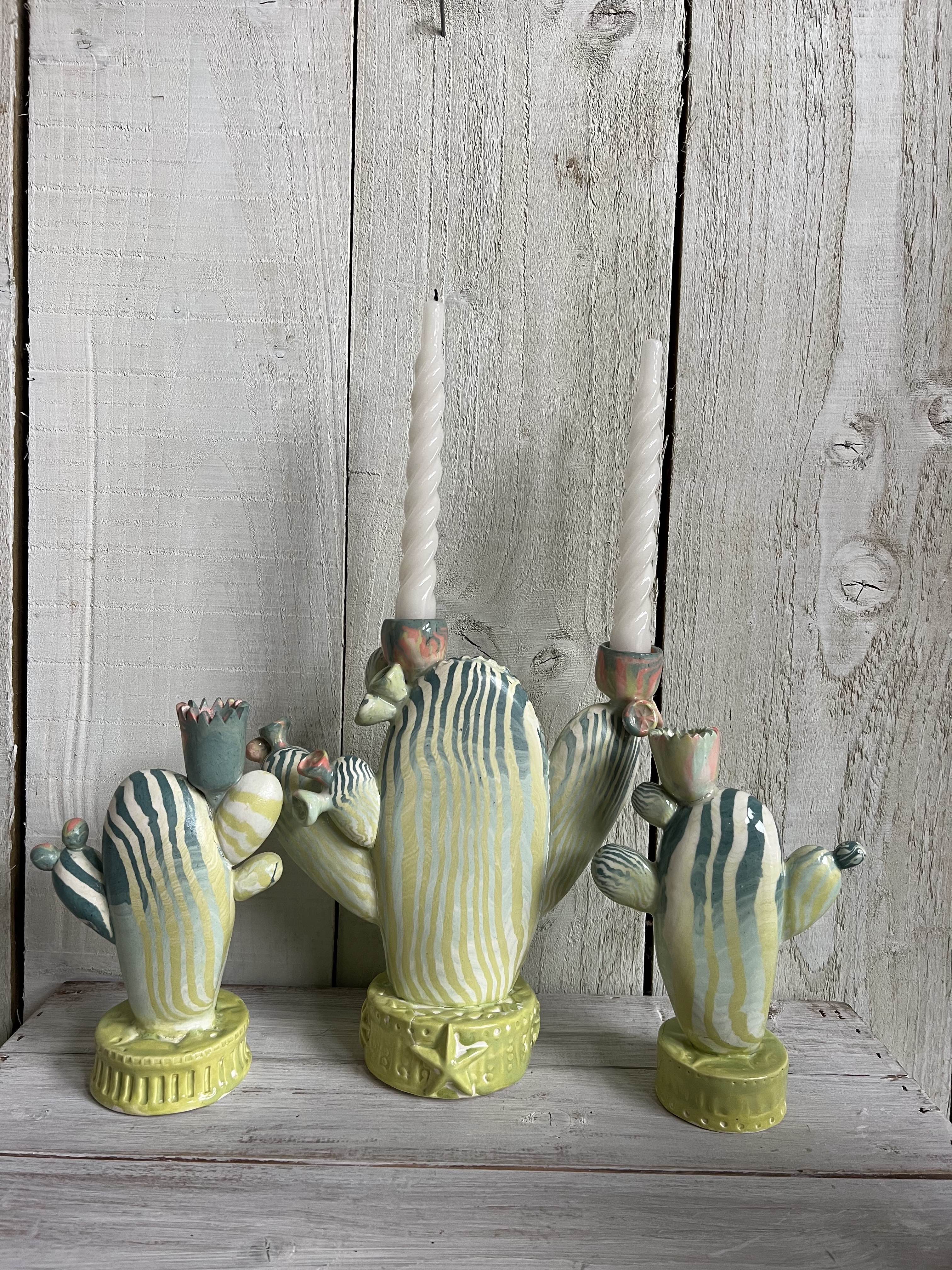 Collection of handmade nerikomi ceramic cactus candle holders with striped clay patterns and taper candles displayed on a rustic wooden shelf.