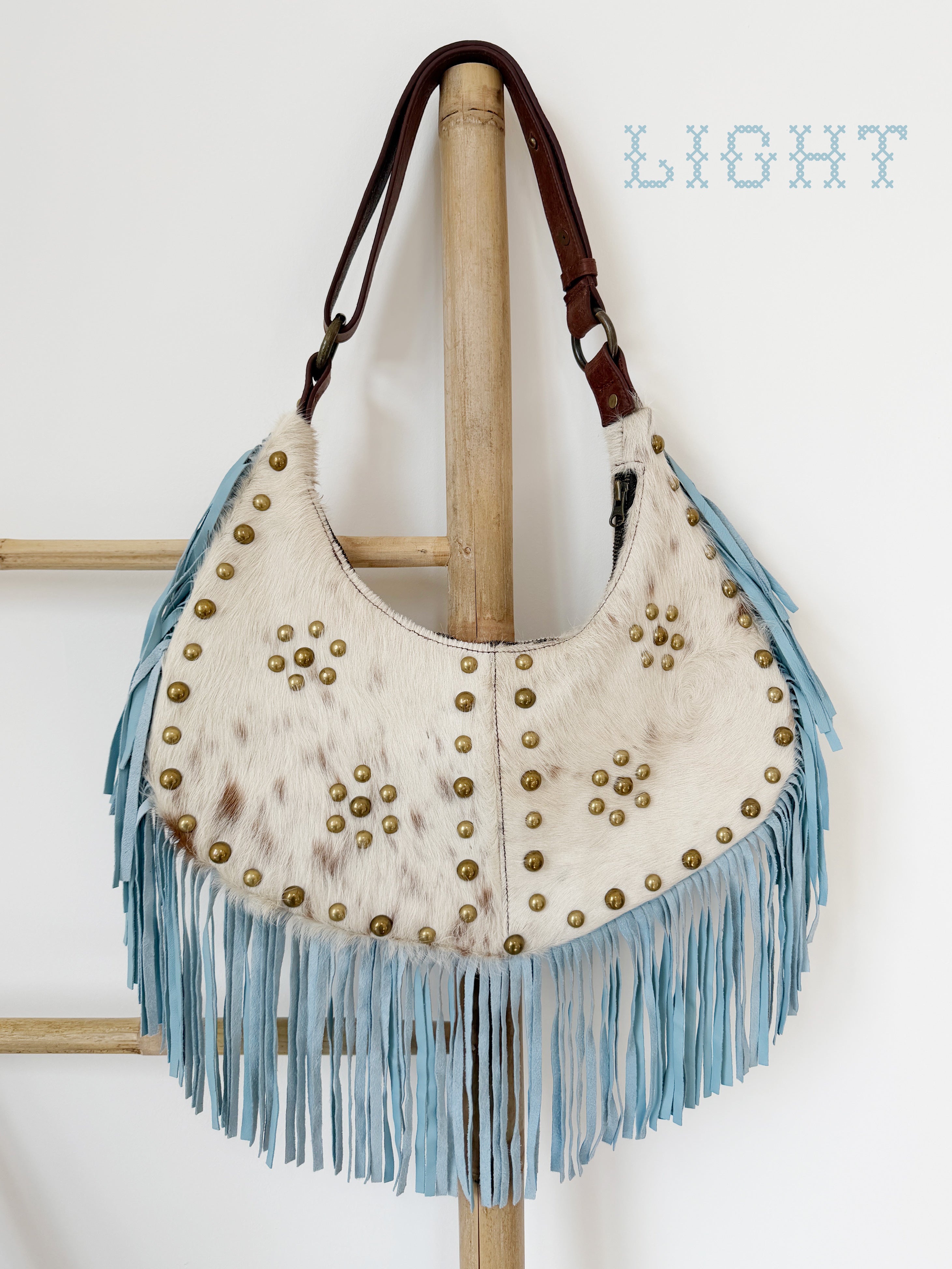 Cream cowhide leather shoulder bag with light blue suede fringe, brass stud detailing, and a brown leather strap.