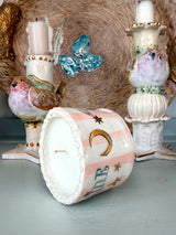 A pastel pink and white striped ceramic candle with gold moon and star details, shown lying on its side on a tabletop. The background features decorative ceramic bird candle holders and a textured brown woven backdrop.