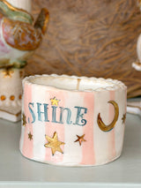 A handmade ceramic candle holder with a cylindrical shape, decorated with pink and white vertical stripes, the word 'SHINE' in blue, gold stars, and a gold crescent moon. It sits on a light gray surface with a warm, textured brown background.