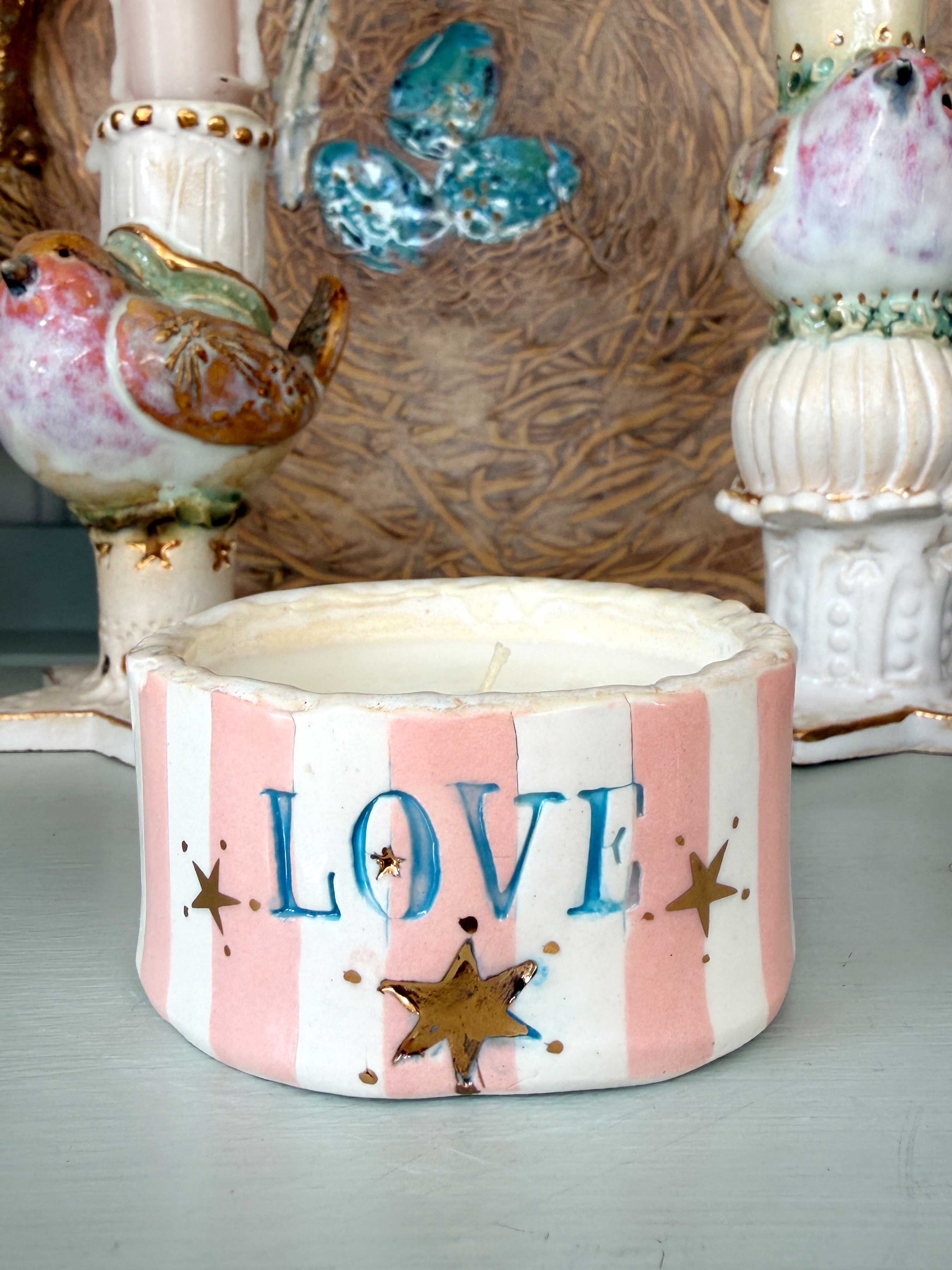 A handmade ceramic candle holder with pink and white vertical stripes, featuring the word 'LOVE' in blue letters and gold star accents, displayed on a tabletop with other decorative items in the background.