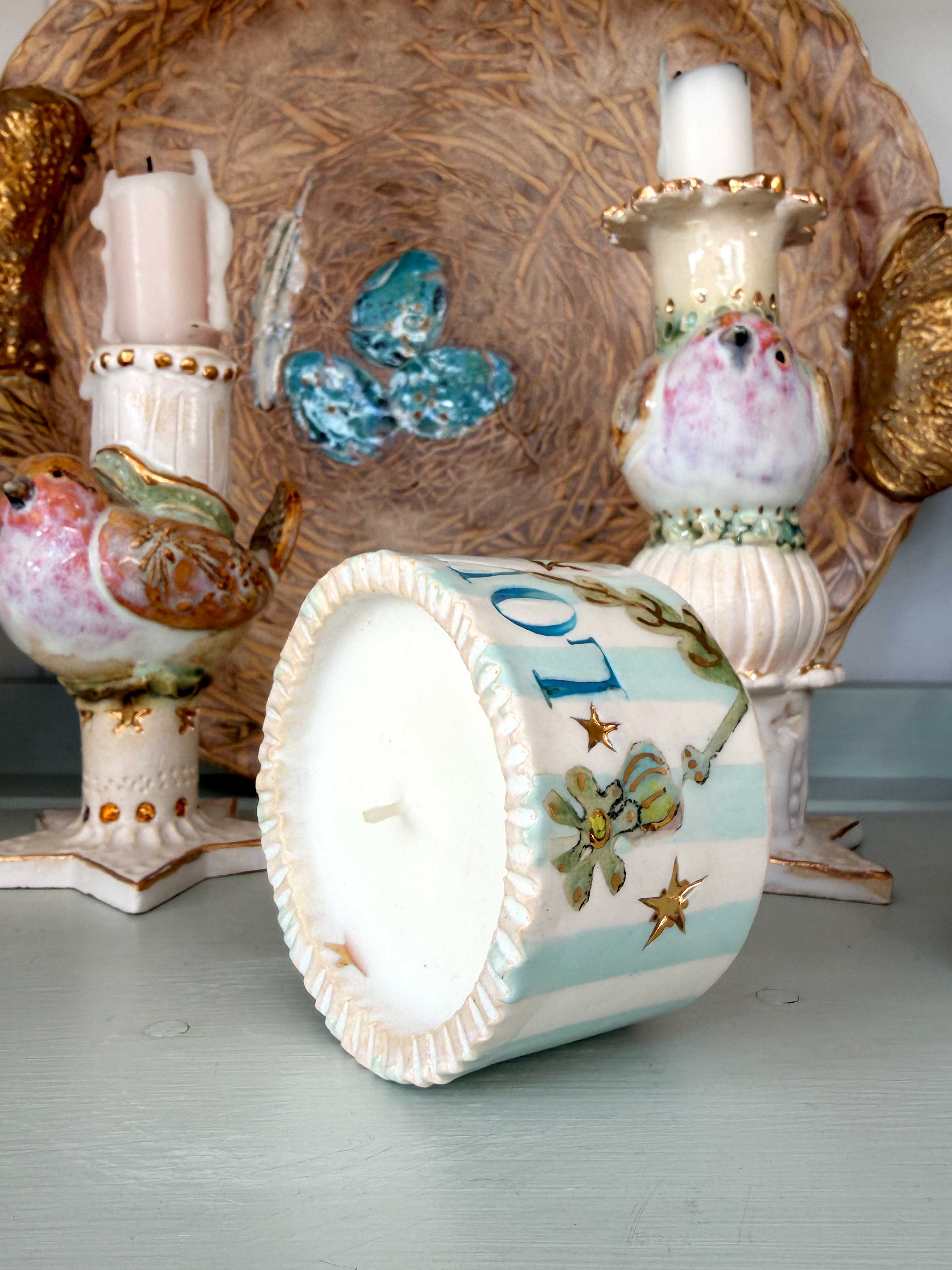 A decorative ceramic candle holder shaped like a bird, shown with one candle inserted and the front of the vessel turned to display a white wax surface with pale blue stripes, floral motifs, and gold star details. Other matching bird-shaped candle holders and a woven tray are visible in the background.