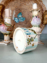 A decorative ceramic candle holder shaped like a bird, shown with one candle inserted and the front of the vessel turned to display a white wax surface with pale blue stripes, floral motifs, and gold star details. Other matching bird-shaped candle holders and a woven tray are visible in the background.