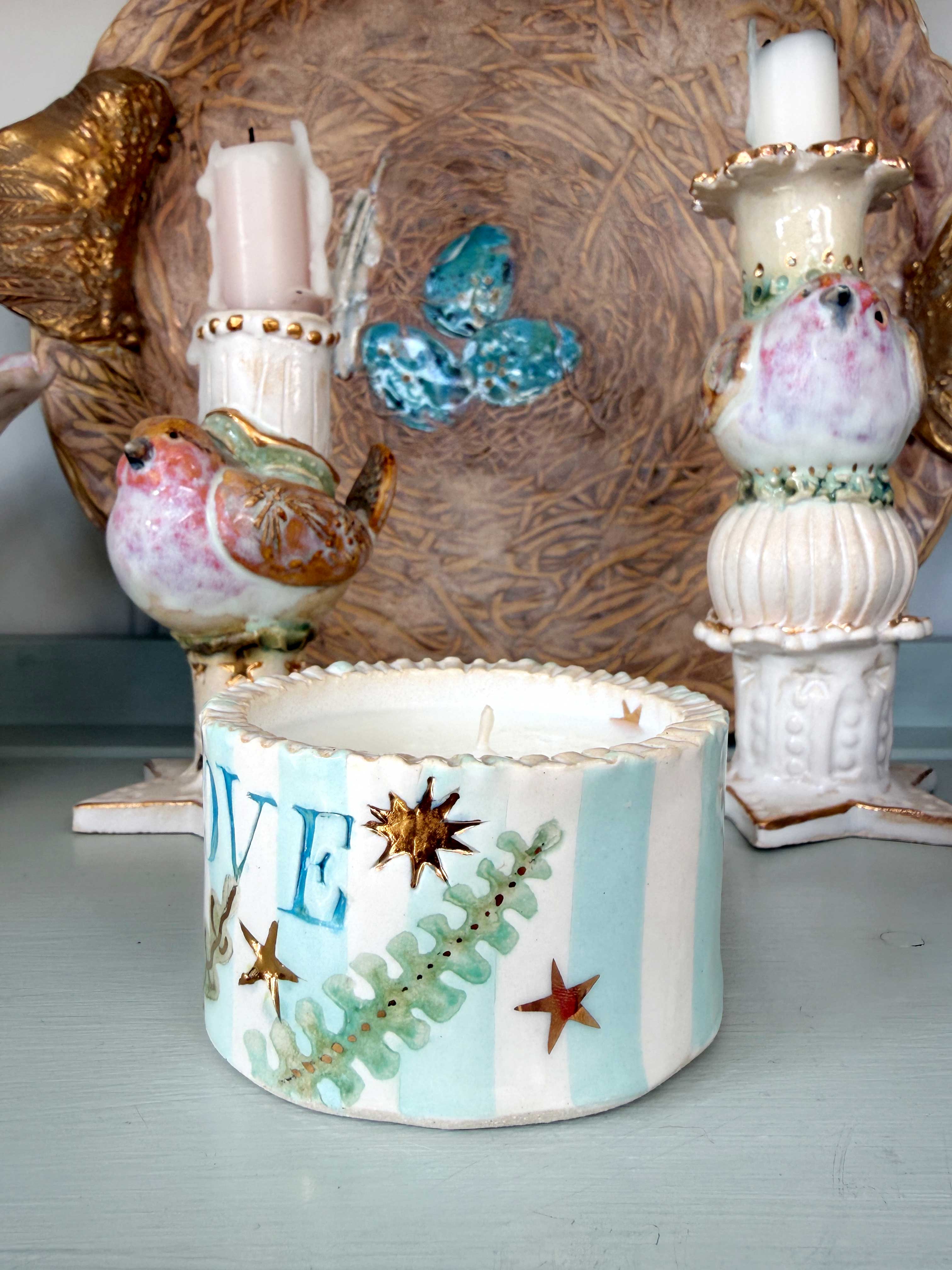 A handmade ceramic candle holder featuring light blue and white stripes, decorated with gold starfish, green fern leaves, and the letter 'E'. It is styled on a light blue surface with other decorative ceramic bird candle holders in the background.