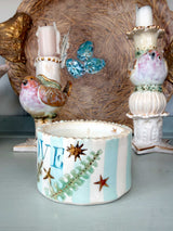 A handmade ceramic candle holder featuring light blue and white stripes, decorated with gold starfish, green fern leaves, and the letter 'E'. It is styled on a light blue surface with other decorative ceramic bird candle holders in the background.