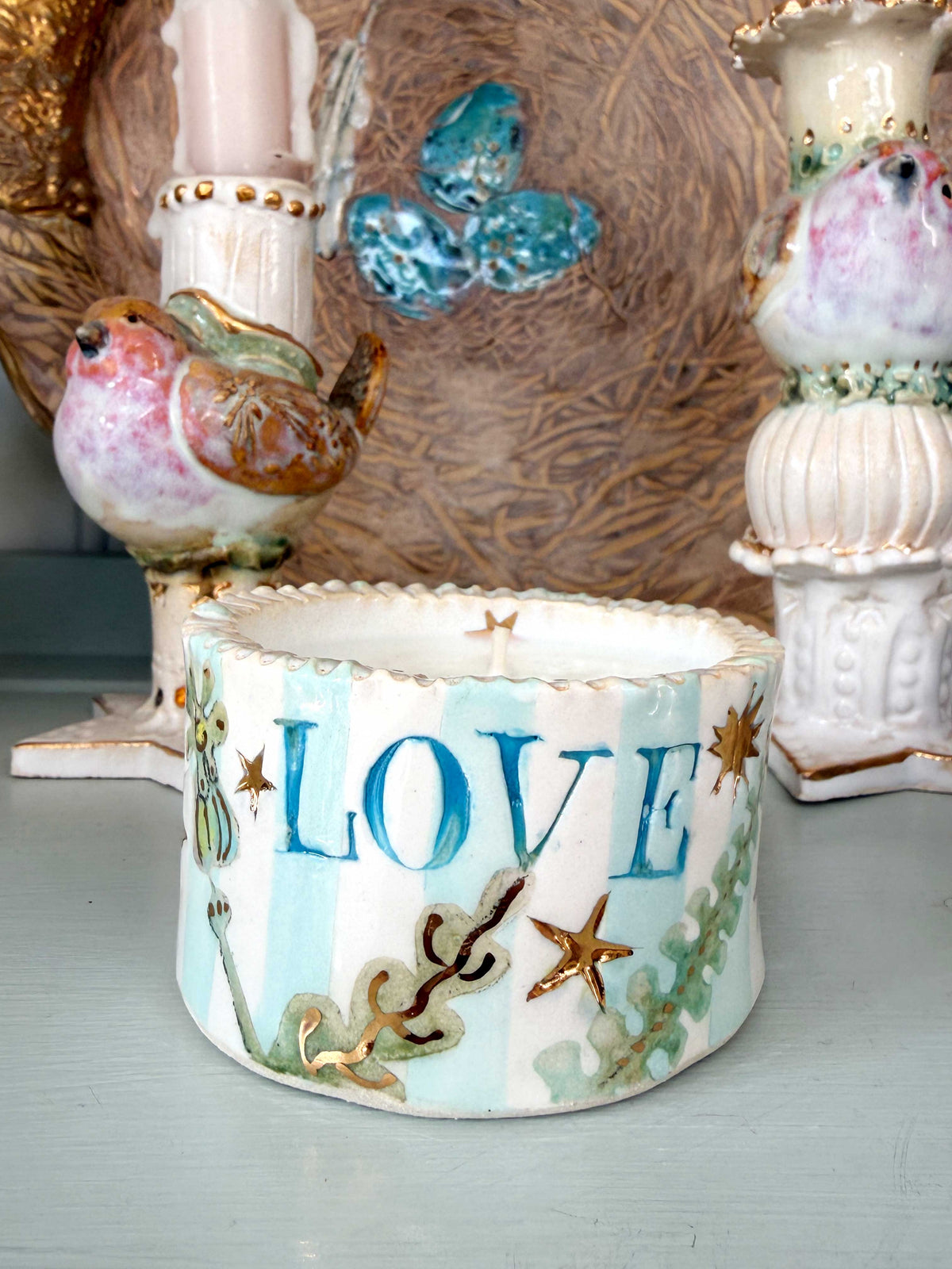 A handmade ceramic candle holder with the word 'LOVE' in blue, decorated with light blue and white stripes, gold starfish, and green seashell motifs, styled on a tabletop with other decorative items.