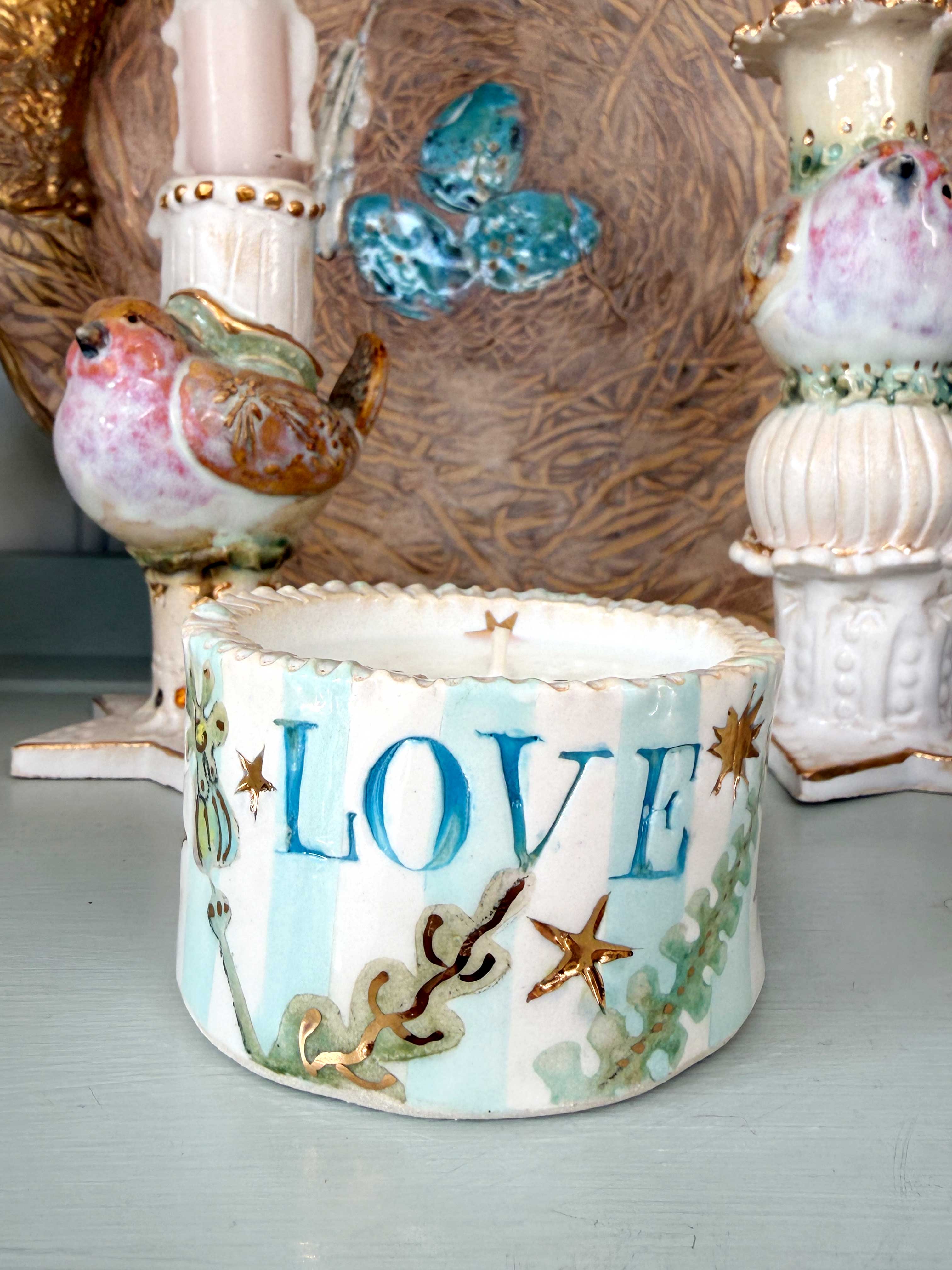 A handmade ceramic candle holder with the word 'LOVE' in blue, decorated with light blue and white stripes, gold starfish, and green seashell motifs, styled on a tabletop with other decorative items.