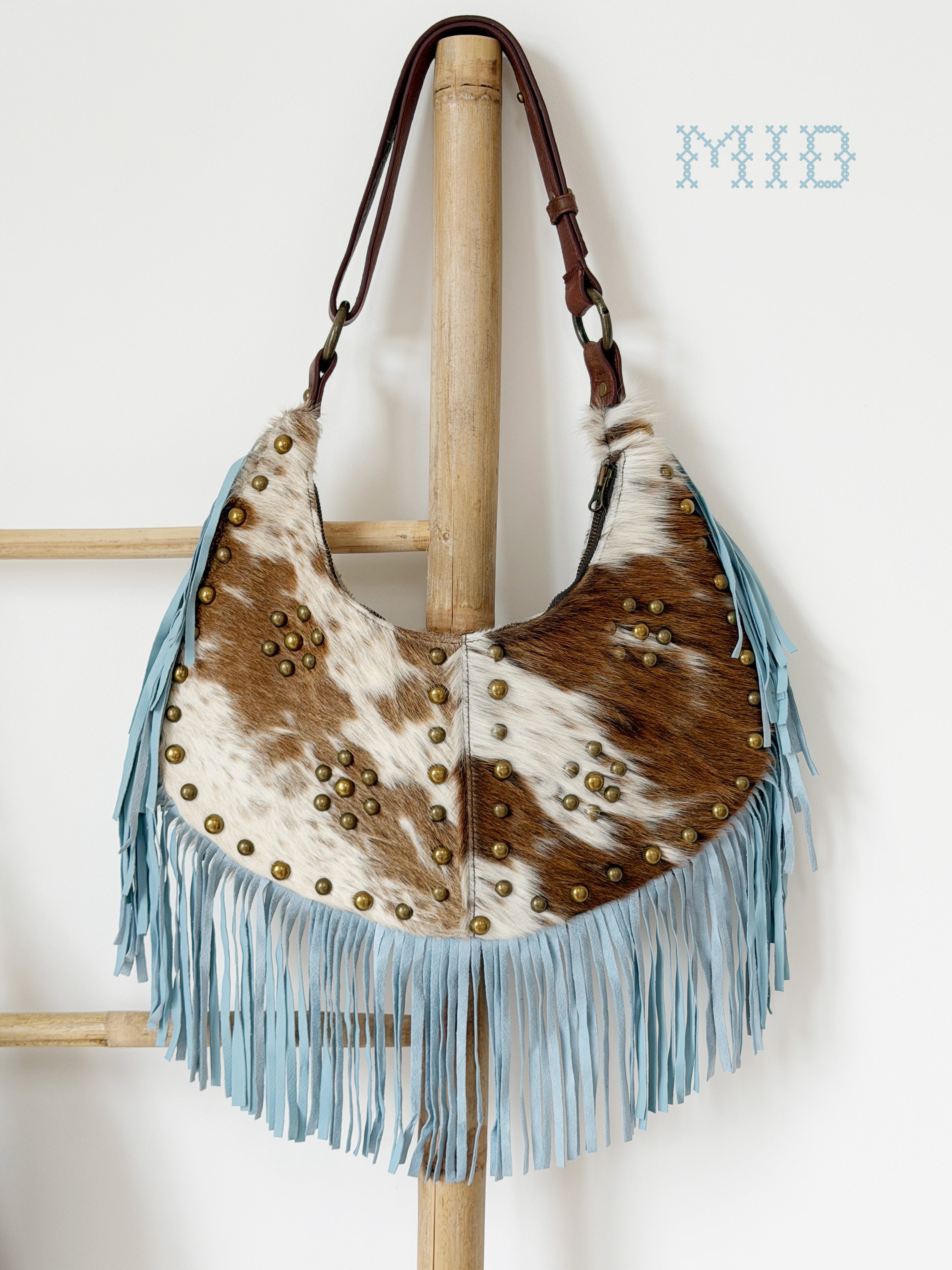 Cream and brown cowhide leather shoulder bag with light blue suede fringe, brass stud detailing, and a brown leather strap.