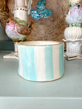 A handmade ceramic candle holder with vertical light blue and white stripes, displayed on a light blue shelf among other decorative ceramic items.