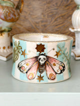 A decorative ceramic candle holder with a painted moth design, featuring pastel blue and cream stripes, pink wings, and gold stars, shown on a tabletop with blurred background decor.