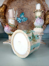 A decorative ceramic bird-shaped candle holder shown tipped on its side, revealing a white wax insert, with other matching bird and bird-of-paradise style candle holders and a large woven backdrop behind them.