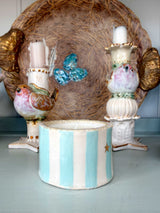 A decorative ceramic bird-themed centerpiece set featuring a cylindrical candle holder with light blue and white vertical stripes in the foreground, flanked by two ornate white bird candlesticks with gold accents, all set against a large woven nest backdrop.