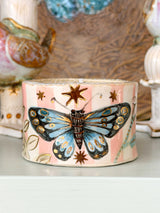 A cylindrical ceramic container or candle holder with a pink and cream glaze, decorated with a large blue and black butterfly and gold stars, displayed on a shelf.