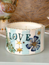 A handmade ceramic candle holder with a scalloped rim, decorated with light blue and white stripes, gold stars, a blue flower, and the word 'LOVE' in dark green. It is shown sitting on a light gray surface with a blurred rustic brown background.