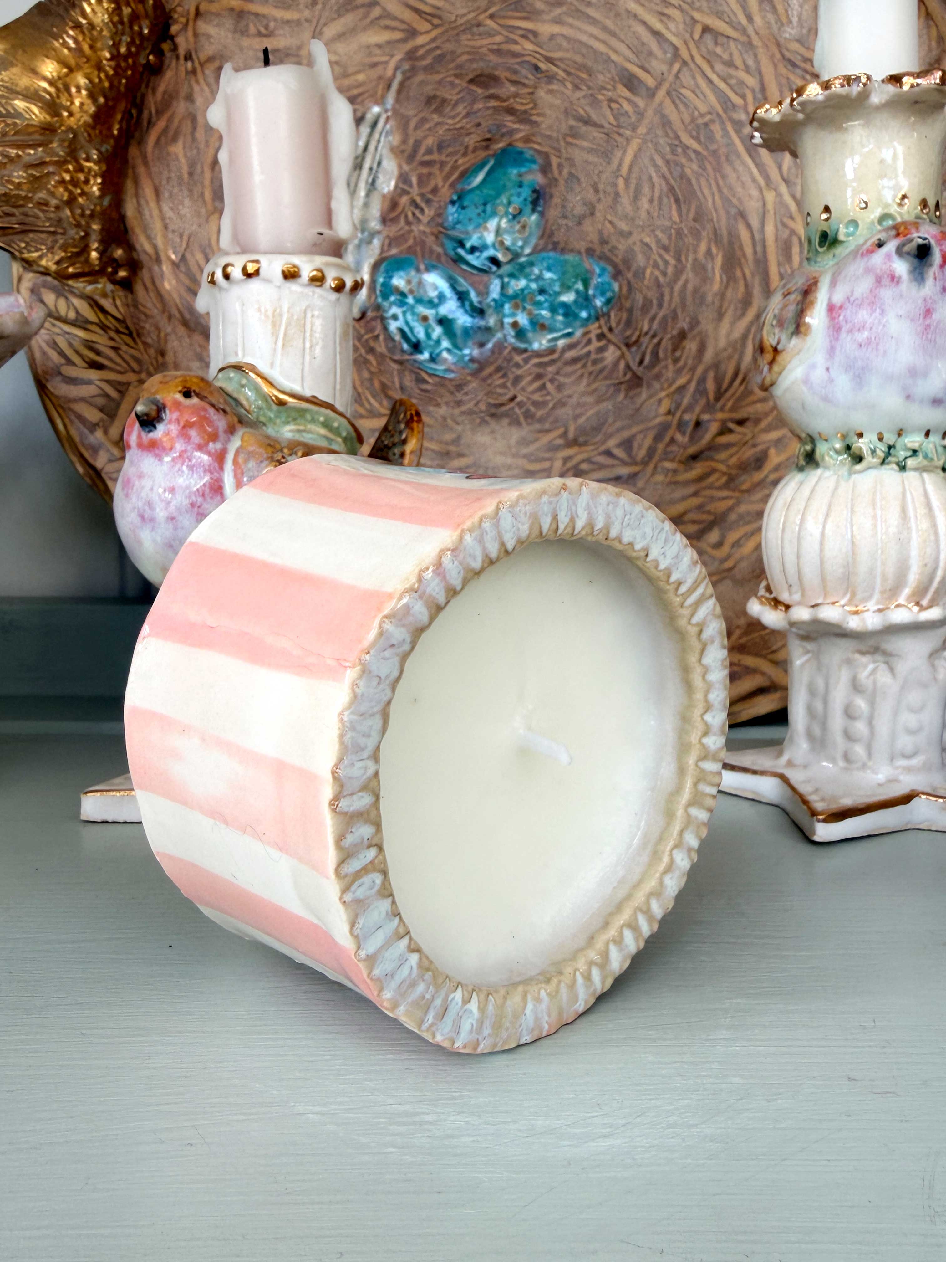 A pastel pink and white striped ceramic candle holder with a scalloped rim, shown tipped on its side to reveal a white candle inside. The background features other decorative ceramic items and a textured brown woven basket.