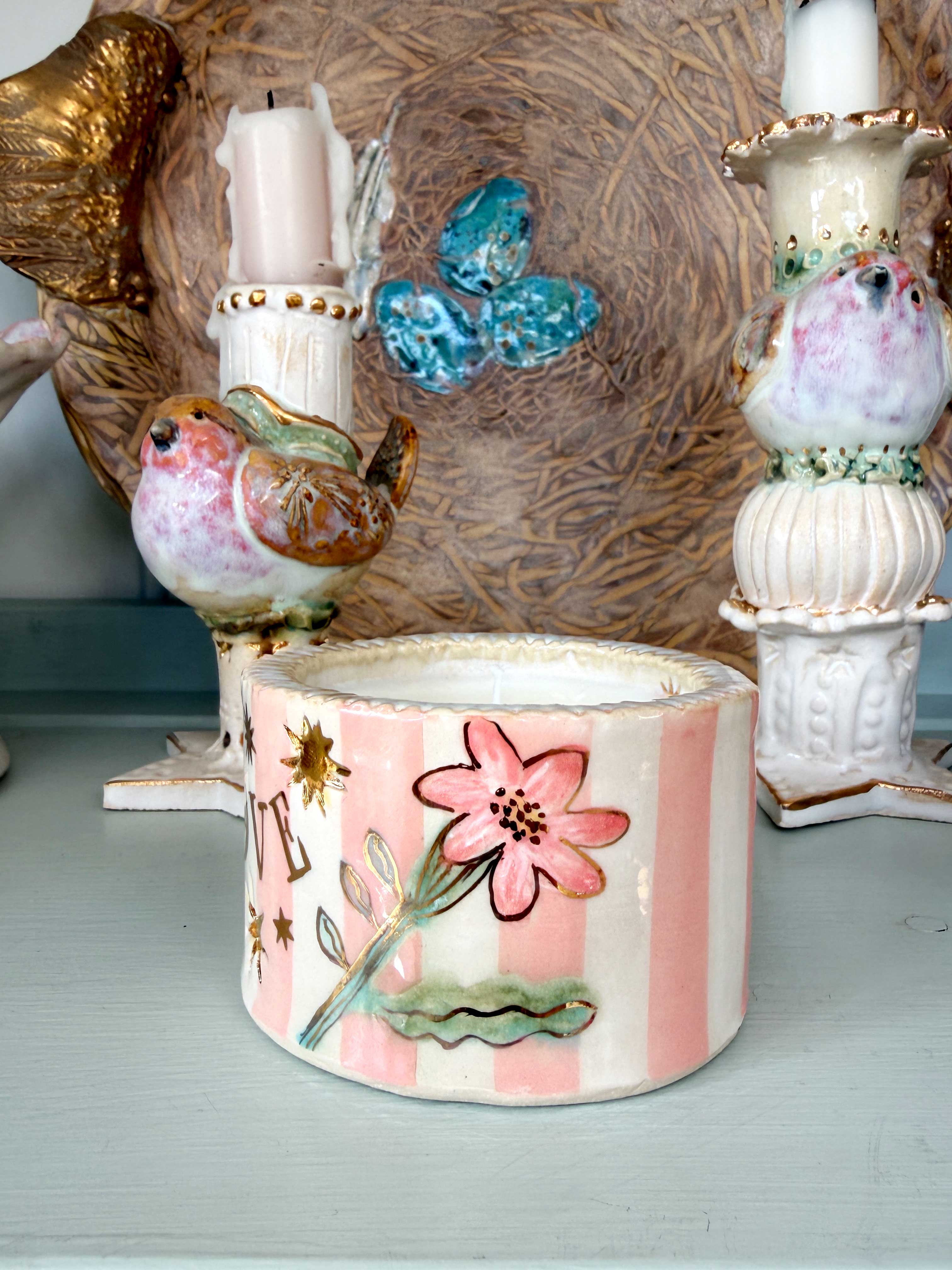 A decorative ceramic candle holder with pink and white vertical stripes, a pink flower motif, and gold stars, displayed on a shelf alongside other ornate ceramic candlesticks.