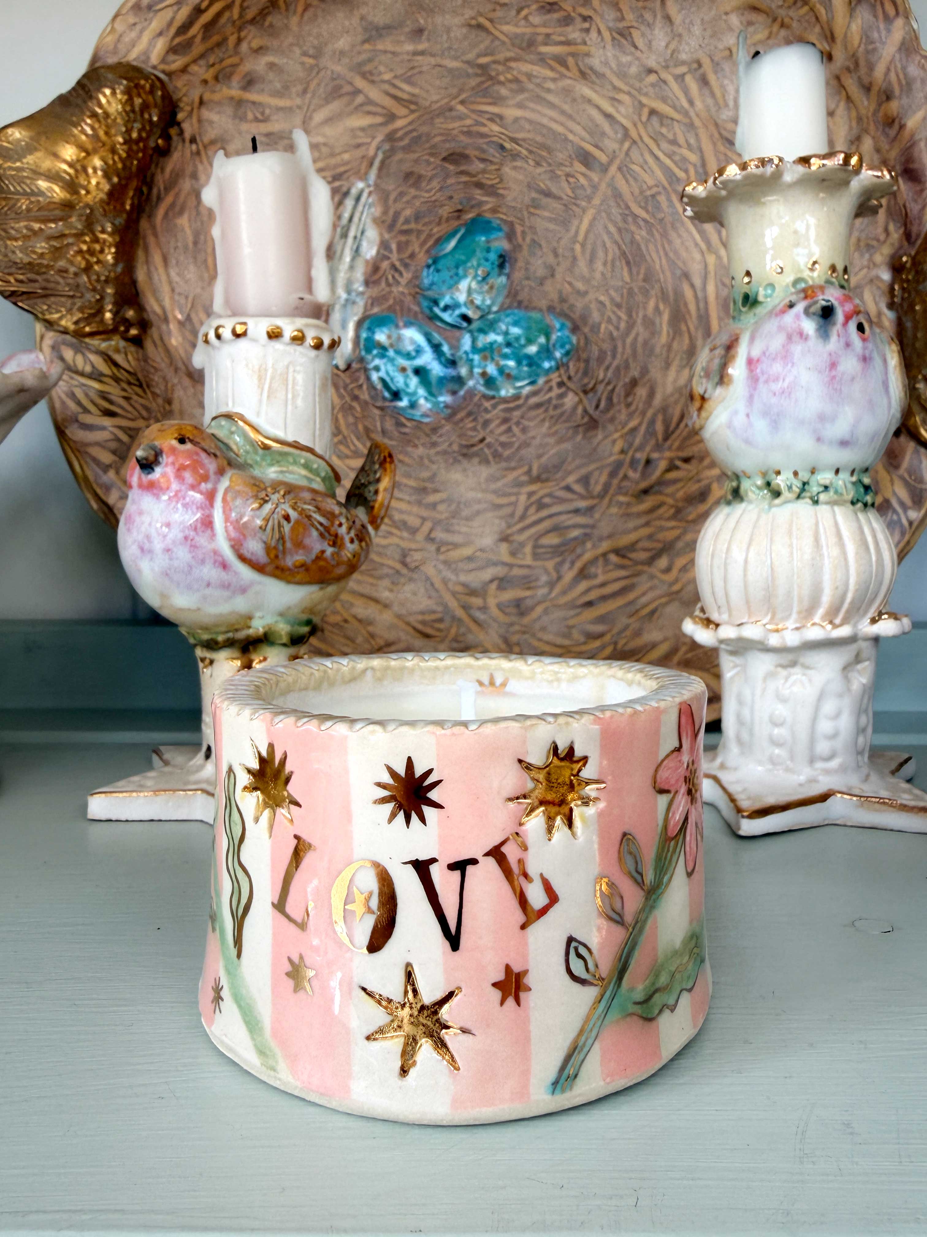 A decorative ceramic candle holder with pink and white vertical stripes, a pink flower motif, and gold stars, displayed on a shelf alongside other ornate ceramic candlesticks.
