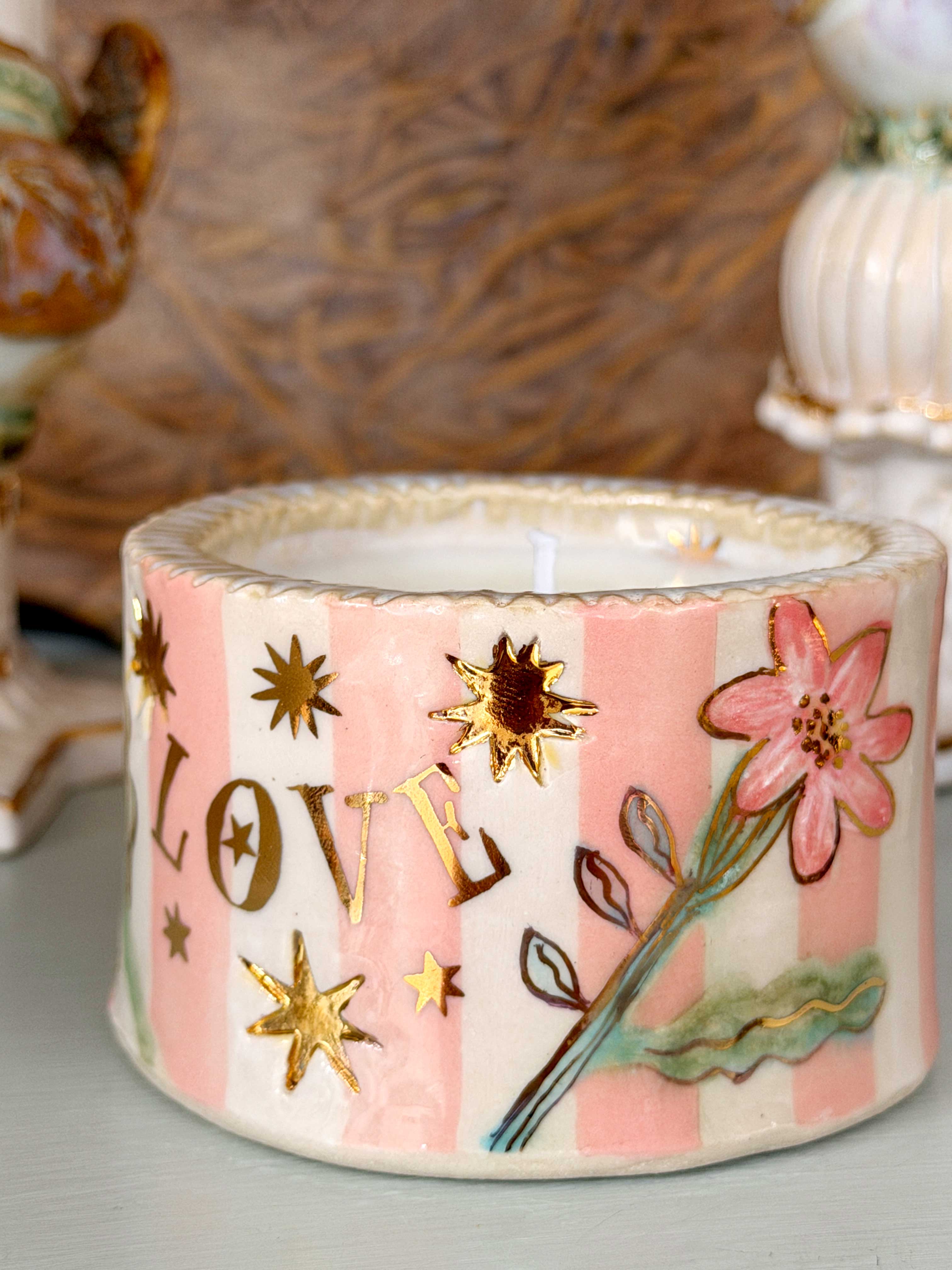 A decorative ceramic candle holder with pink and white vertical stripes, a pink flower motif, and gold stars, displayed on a shelf alongside other ornate ceramic candlesticks.