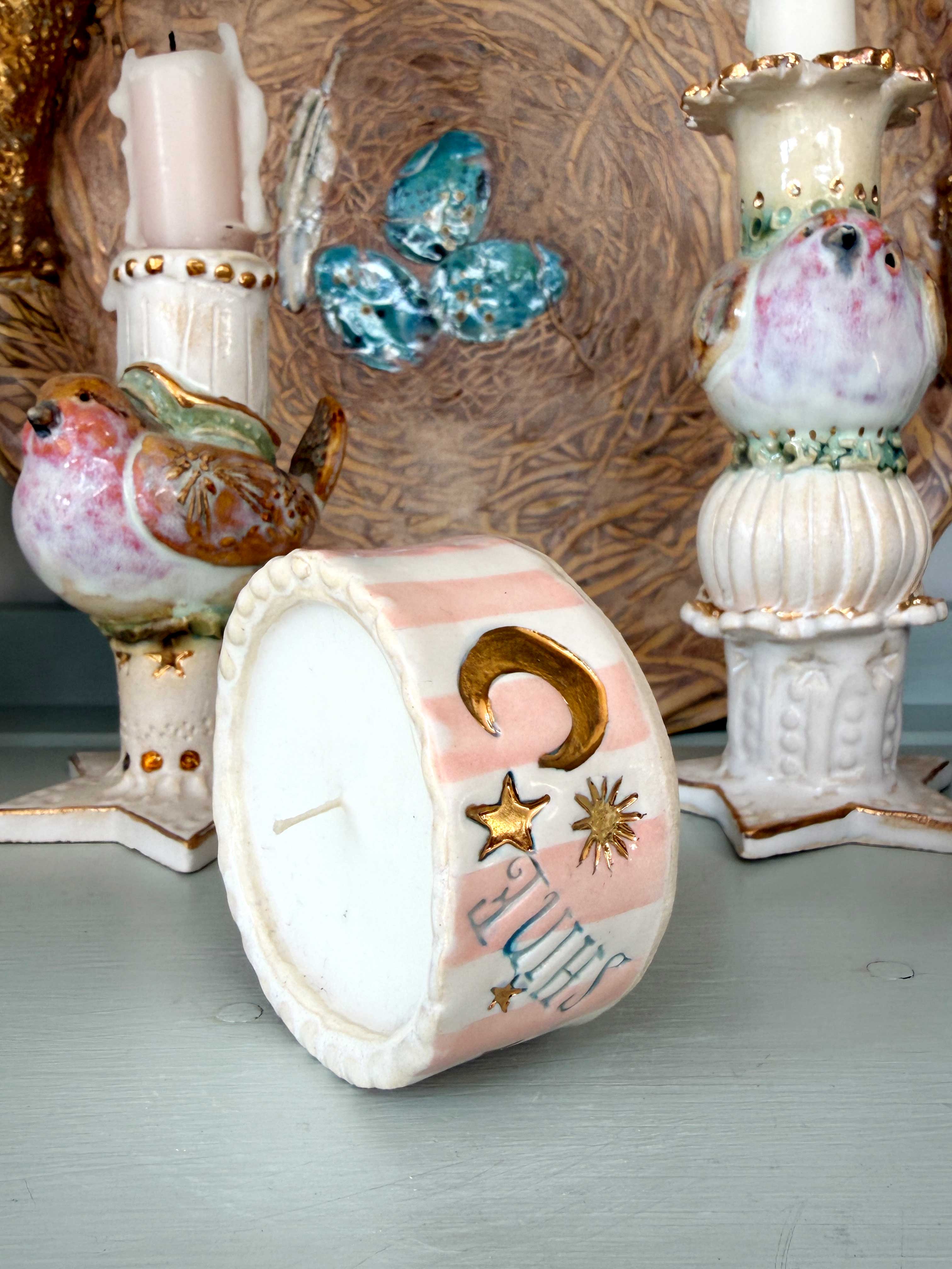 A small decorative candle holder shaped like a drum, shown lying on its side. The drum is white with pastel pink stripes, gold crescent moon and star motifs, and a blue-and-white hand-drawn bird design.