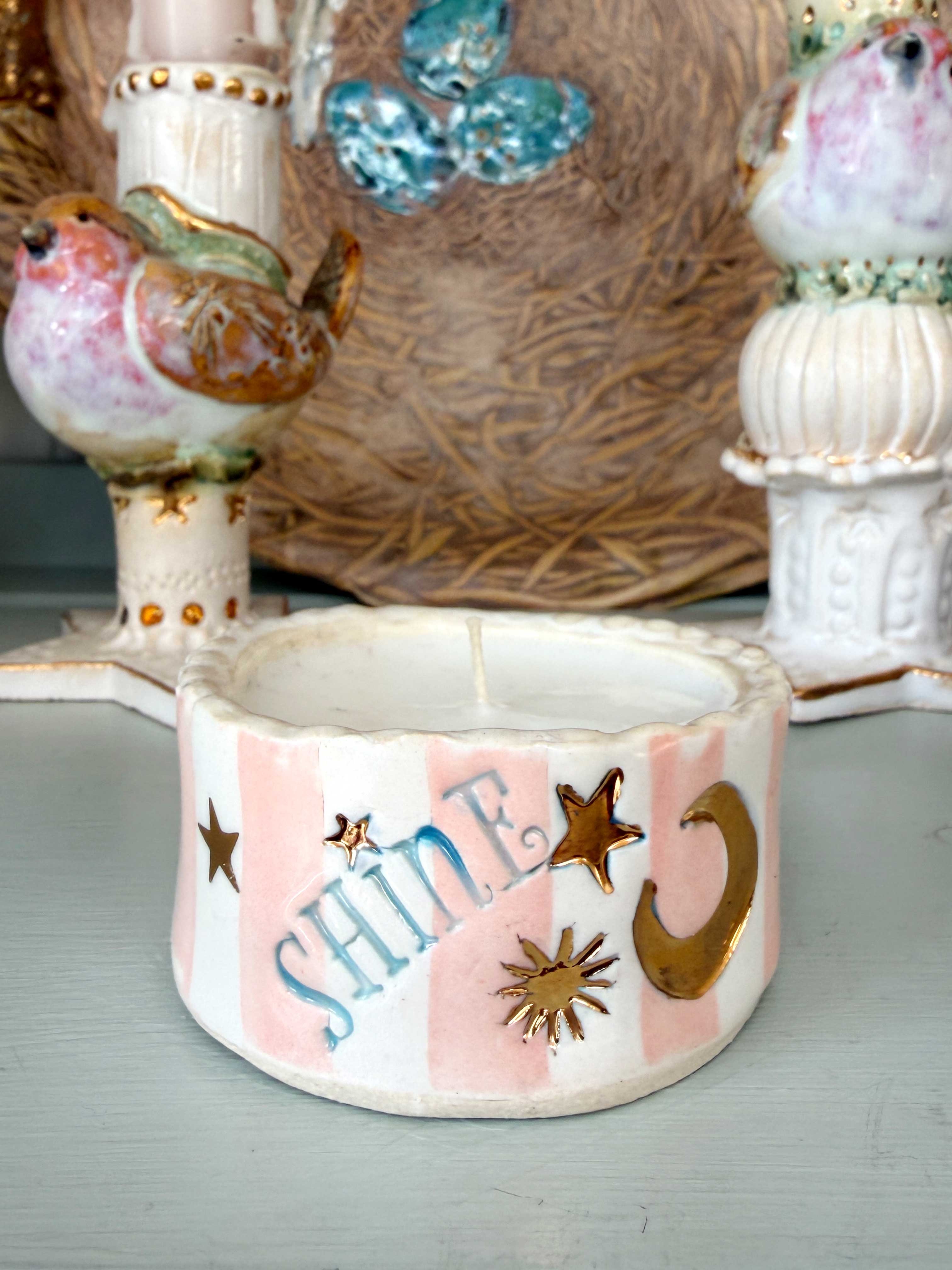A small ceramic candle holder with a white and light pink striped design, decorated with gold stars, a crescent moon, and the word 'SHINE' in blue script. The candle holder is shown in a styled tabletop setting with other decorative ceramic items in the background.