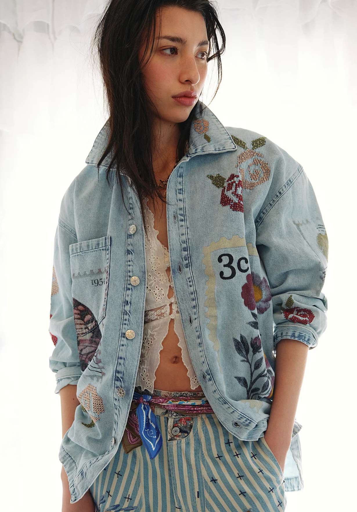 Front view of a person wearing a light wash denim button-up shirt featuring vintage-inspired postage stamp patchwork with floral cross-stitch artwork, paired with blue and white striped pants.