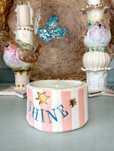 A handmade ceramic candle holder with pink and white vertical stripes, decorated with gold stars, blue lettering spelling 'SHINE', and a white wax candle placed inside. The candle is styled on a light blue-gray surface with decorative bird and bird-of-paradise ceramic candlesticks and a woven basket in the background.