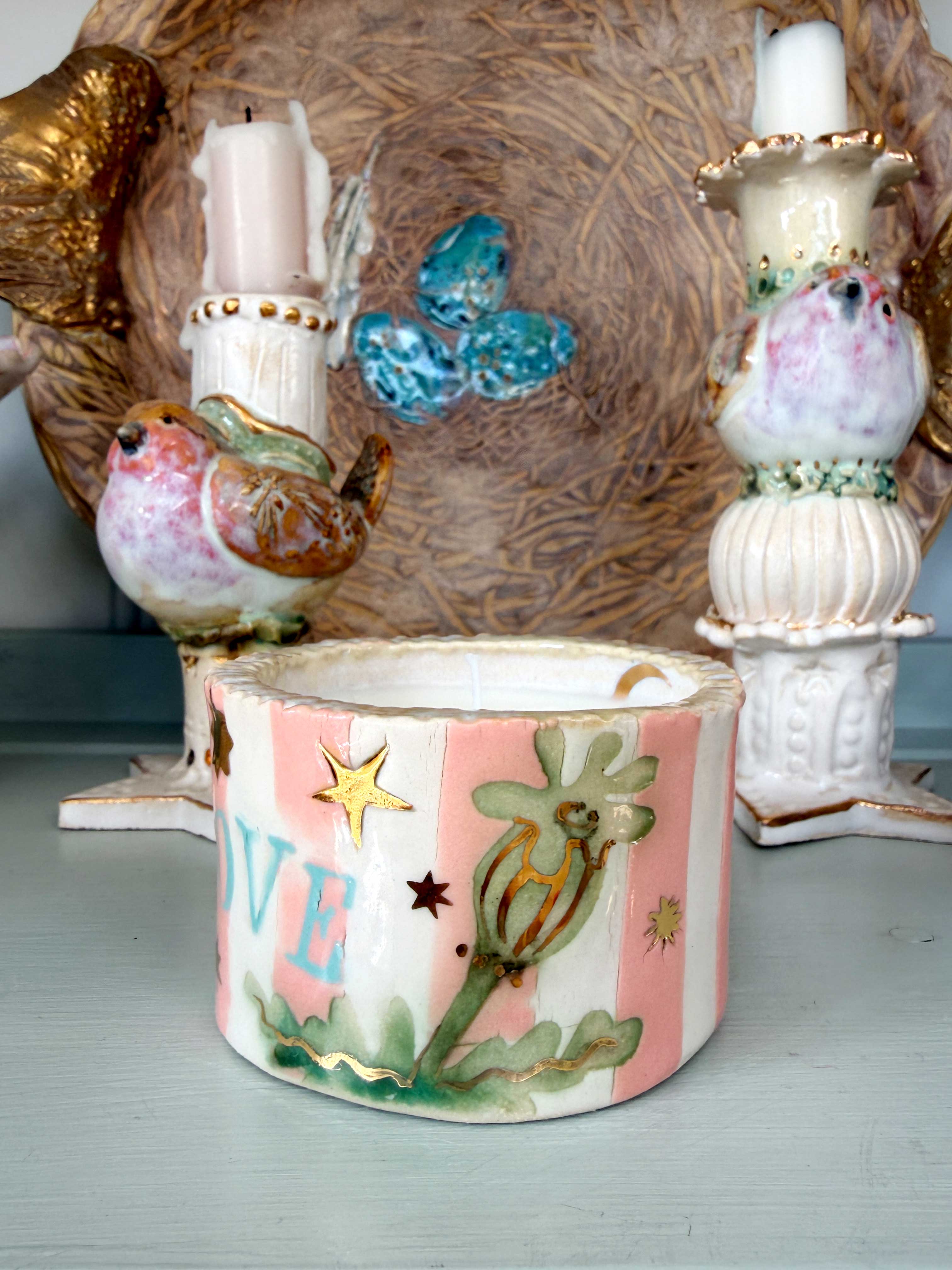 A handmade ceramic candle holder featuring a pink and white striped design with gold glitter stars, a green poppy flower, and the letter 'VE' in blue. The holder is styled on a light blue-gray surface with other decorative ceramic bird and bird-of-paradise figurines in the background.