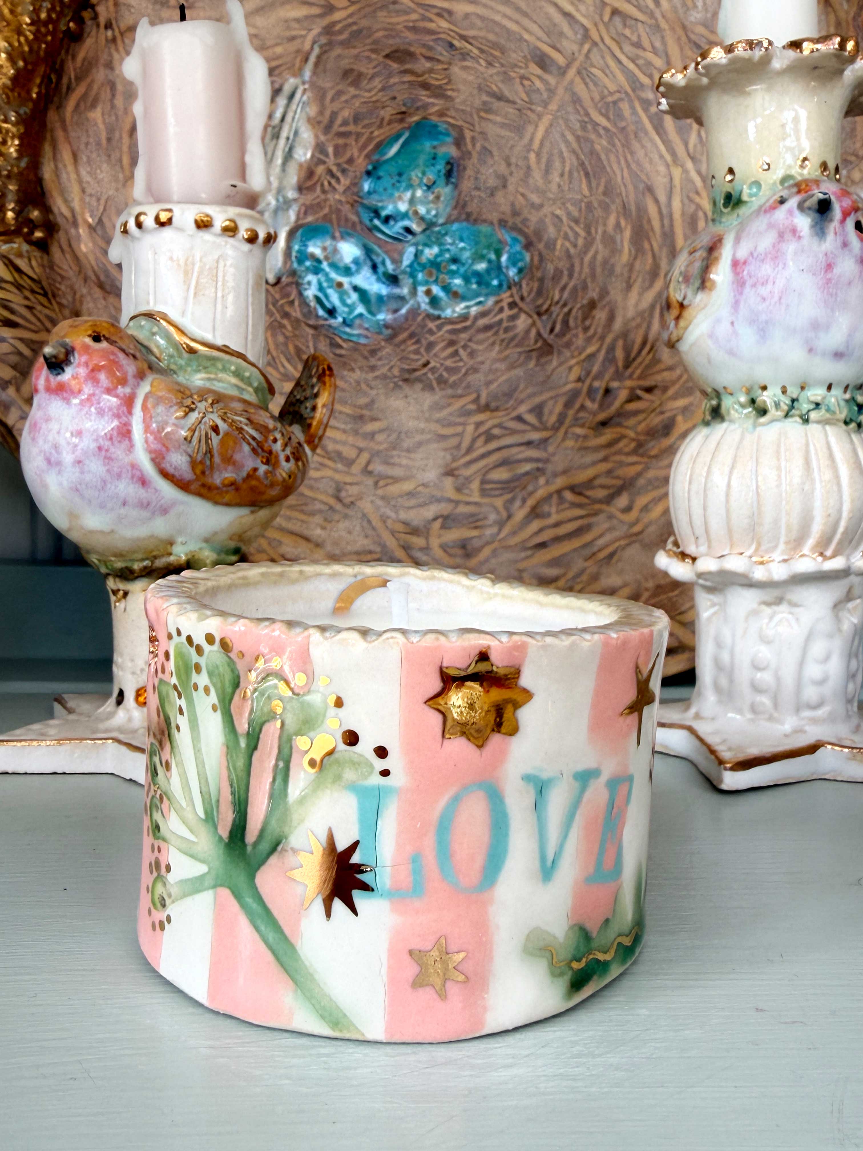 A handmade ceramic candle holder with a pink and white striped pattern, decorated with green leaves, gold stars, and the word 'LOVE' in blue. It is styled on a tabletop with other decorative ceramic items in the background.