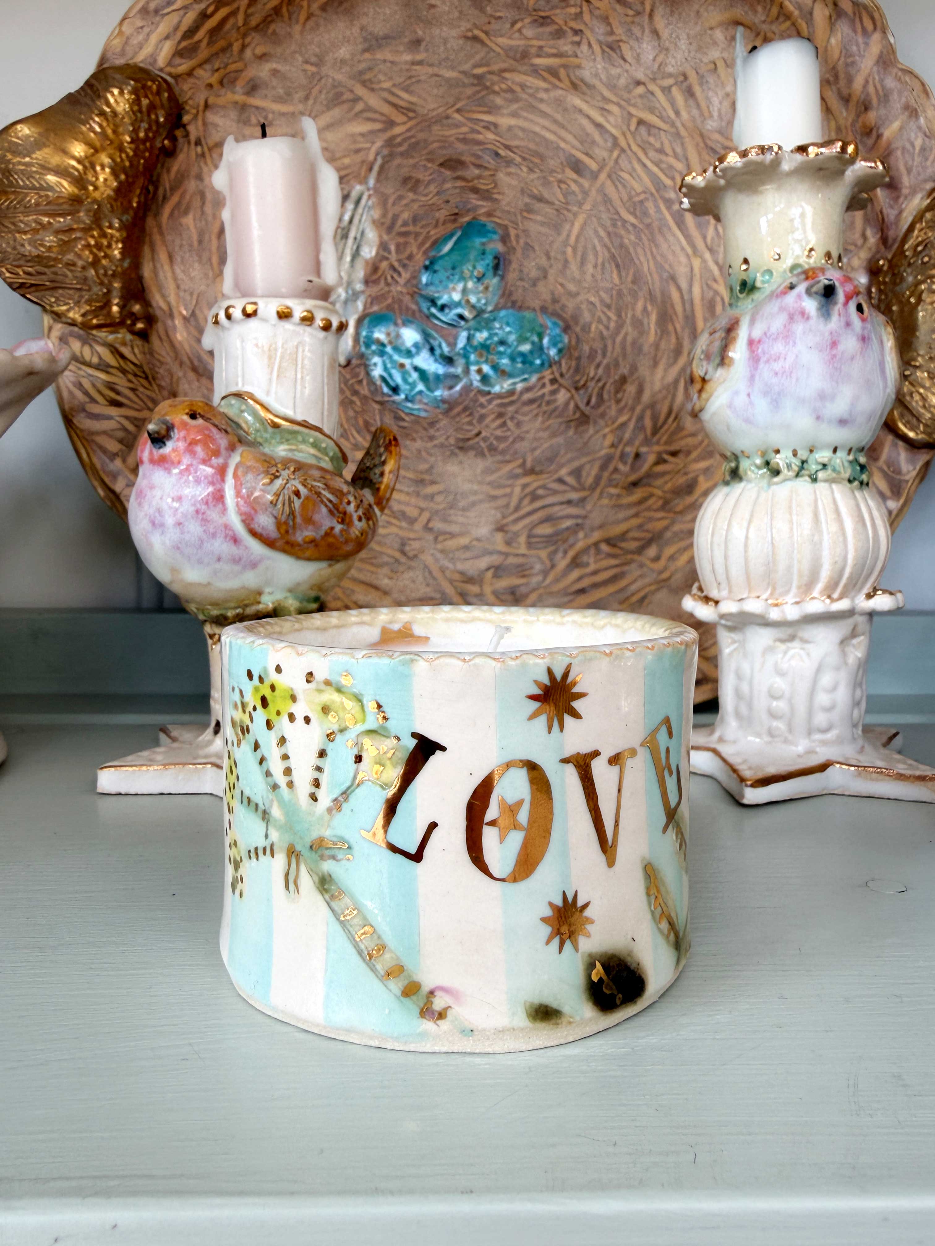 A decorative ceramic candle holder with the word 'LOVE' in gold lettering, decorated with a butterfly, stars, and floral motifs on a light blue and white striped background, displayed on a shelf.