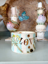 A decorative ceramic candle holder with the word 'LOVE' in gold lettering, decorated with a butterfly, stars, and floral motifs on a light blue and white striped background, displayed on a shelf.