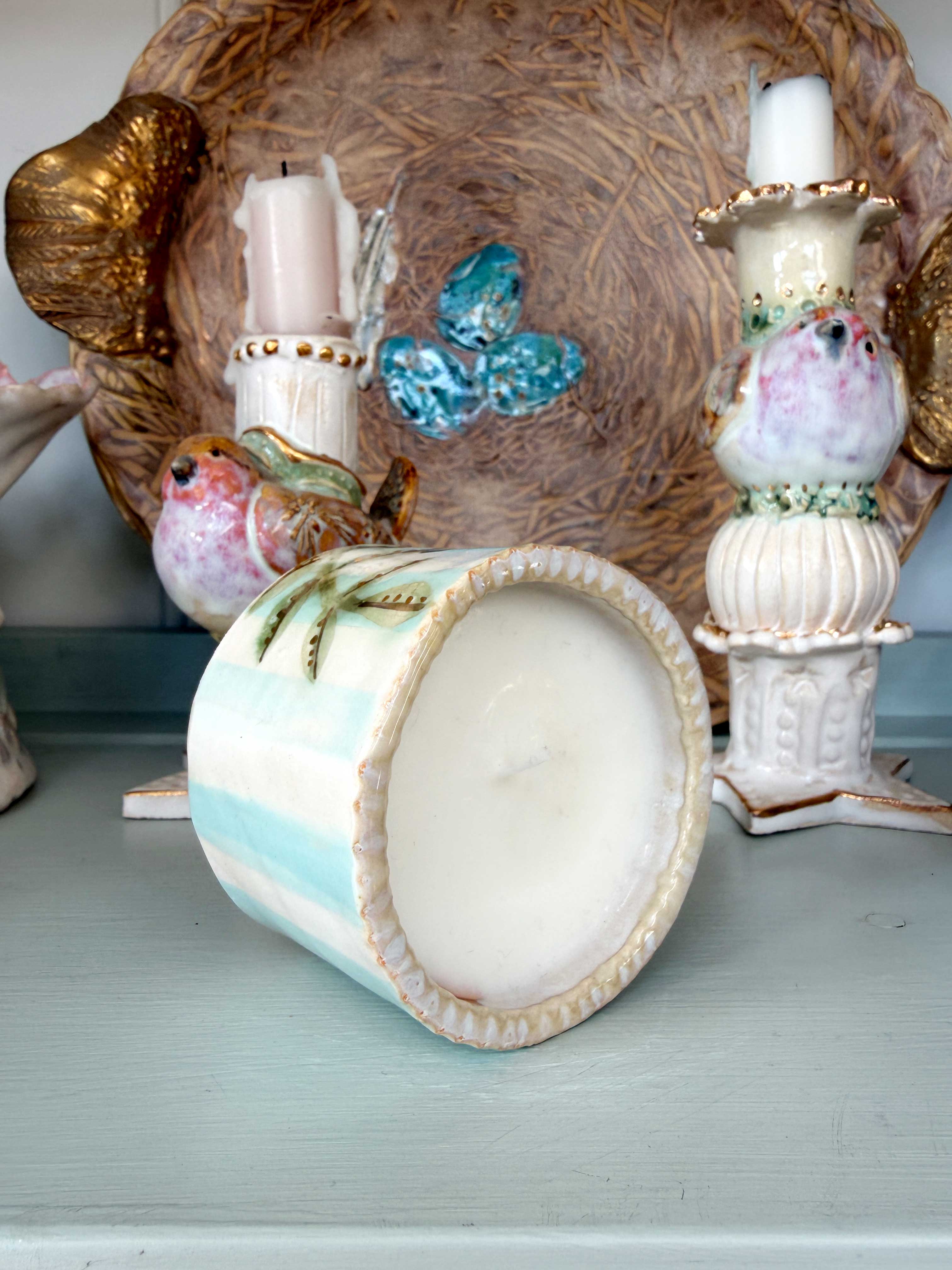 A pastel blue and white striped ceramic candle jar with a scalloped cream rim, shown tipped on its side on a painted surface. The background features vintage-style decorative bird figurines and a large woven basket.