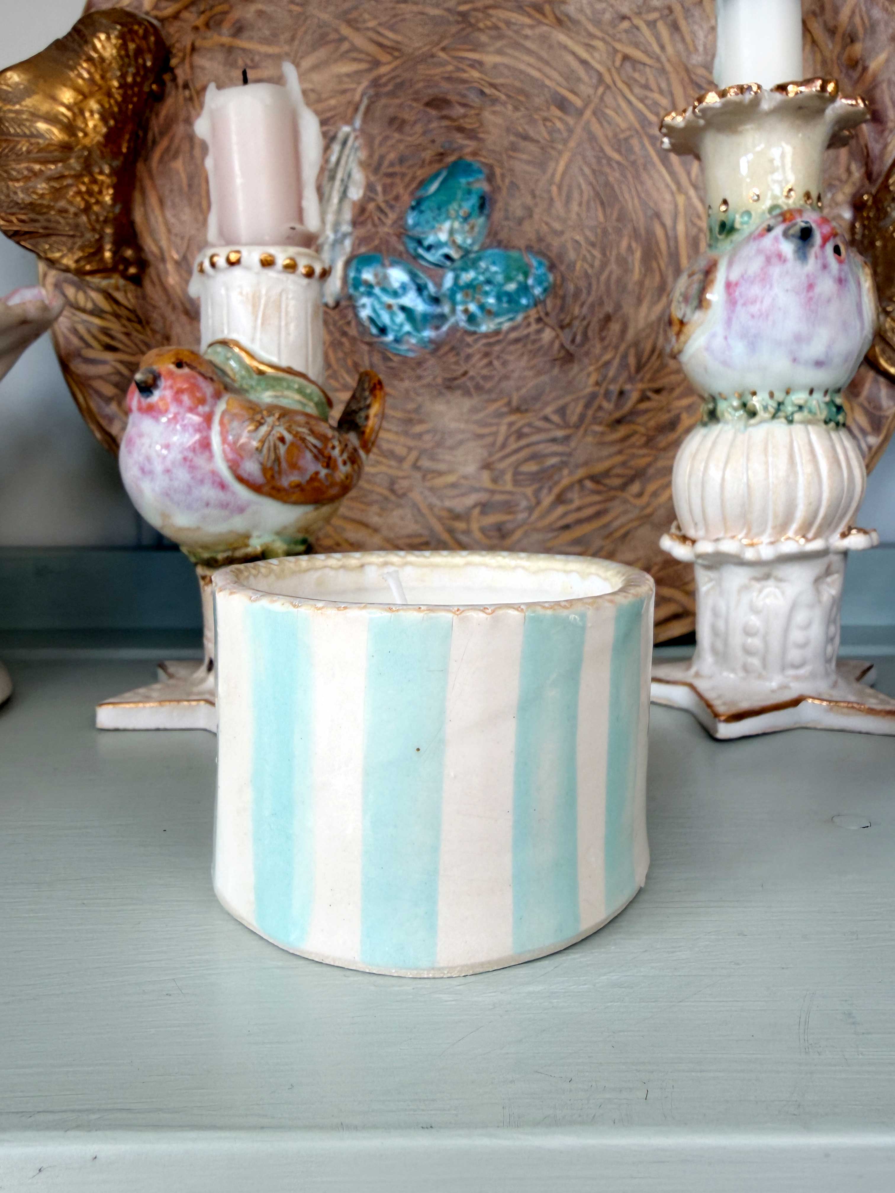 A cylindrical ceramic container with vertical light blue and white stripes, styled on a painted surface with decorative bird figurines and a textured background.