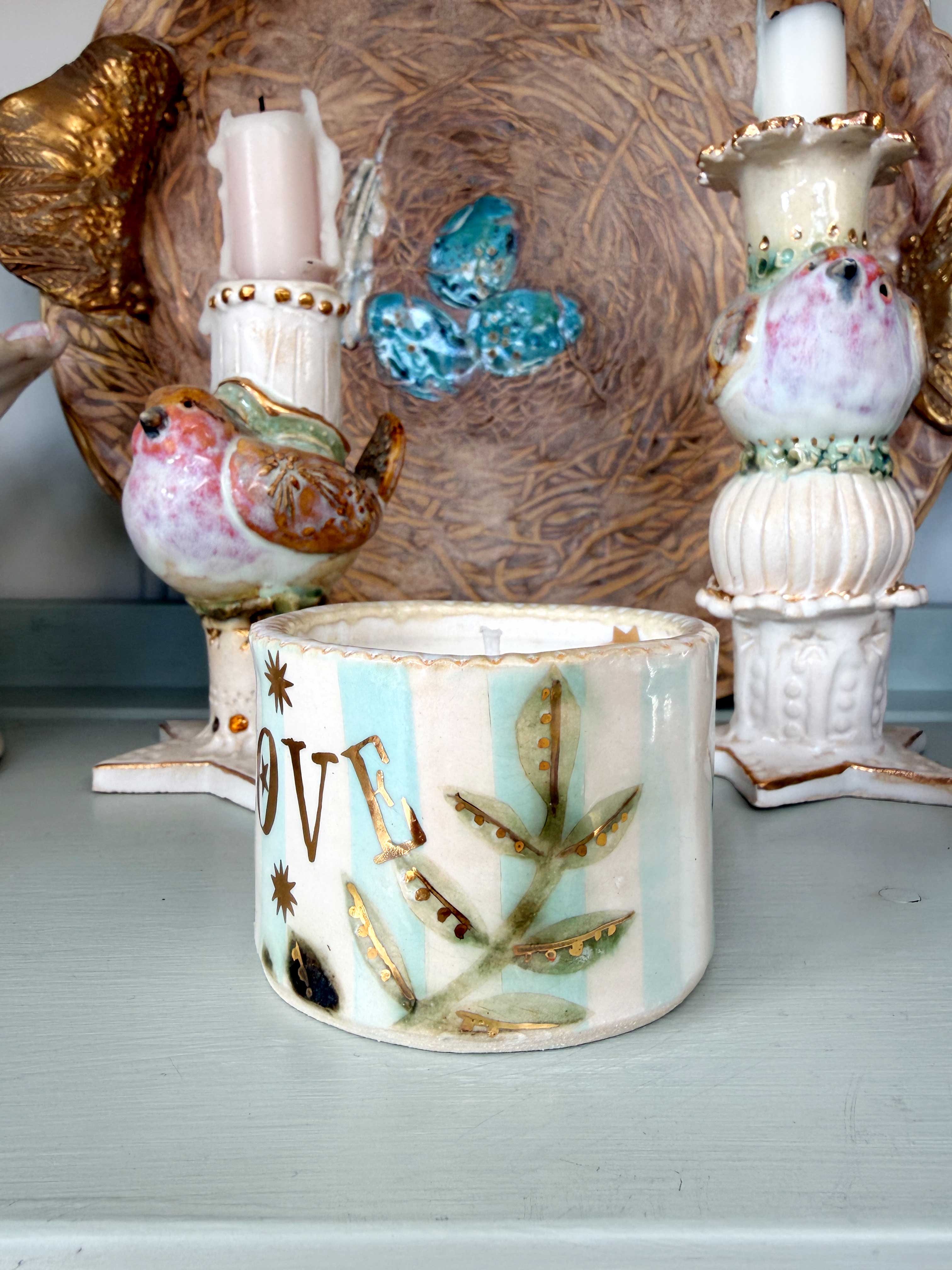 A decorative ceramic candle holder with the word 'LOVE' in gold lettering, decorated with a butterfly, stars, and floral motifs on a light blue and white striped background, displayed on a shelf.