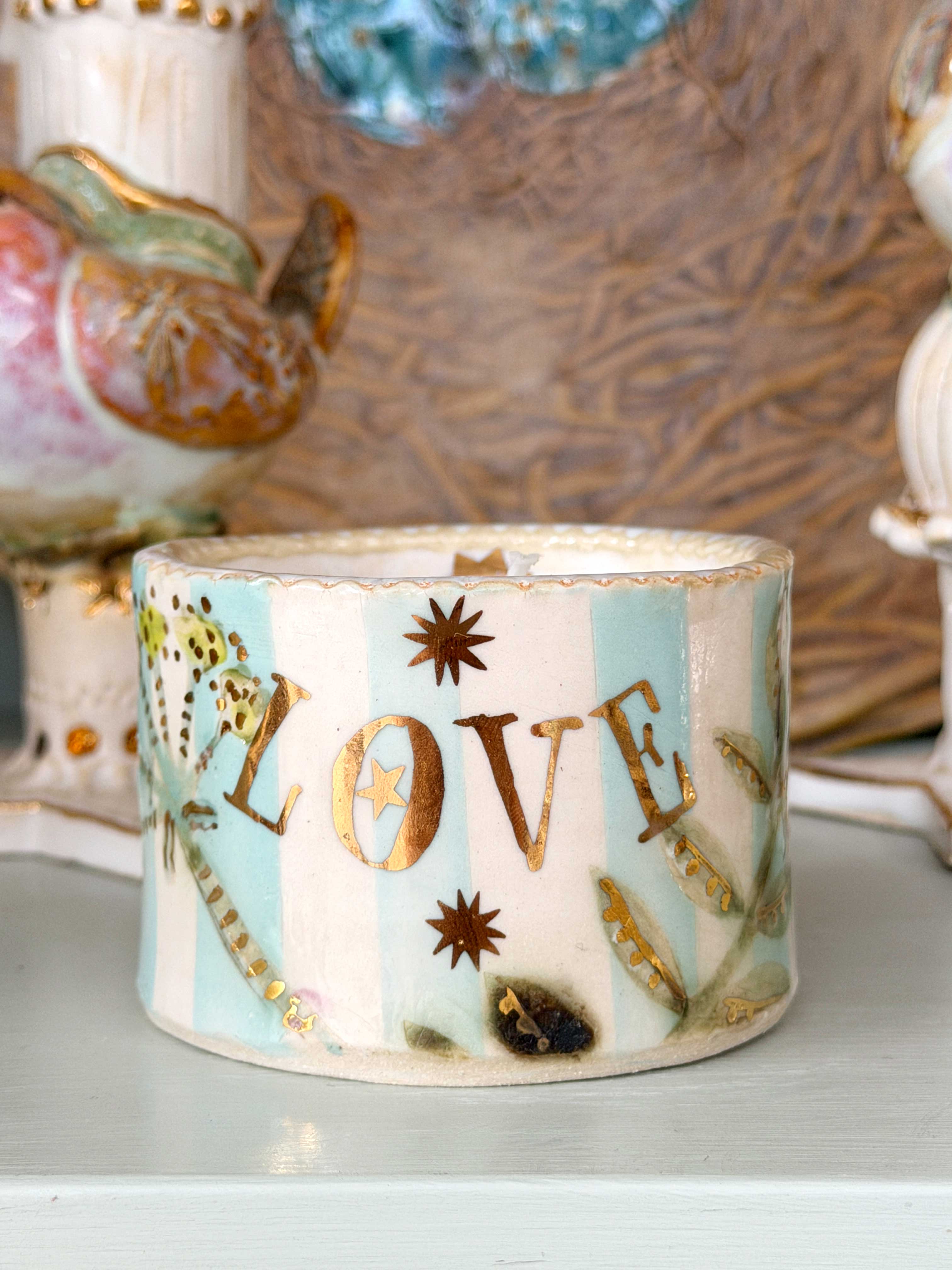 A decorative ceramic candle holder with the word 'LOVE' in gold lettering, decorated with a butterfly, stars, and floral motifs on a light blue and white striped background, displayed on a shelf.