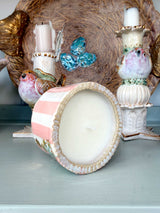 A pink and white striped ceramic candle holder with a scalloped rim, shown tipped over to reveal white wax inside, set against a styled background of decorative ceramic bird candle holders and a gold tray.