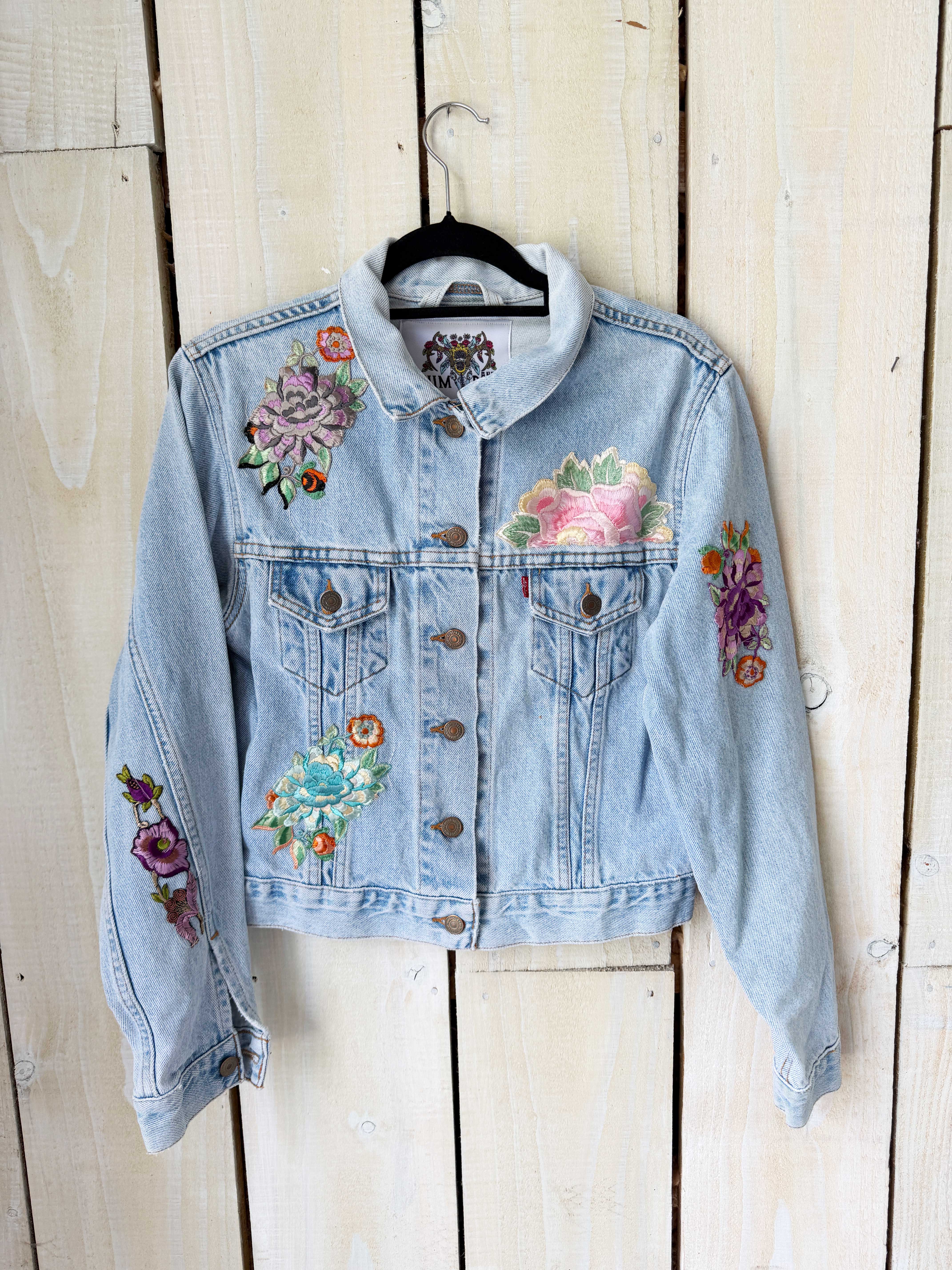 Light blue denim jacket with colourful vintage textile patchwork on the front. hanging on a rustic white wooden wall