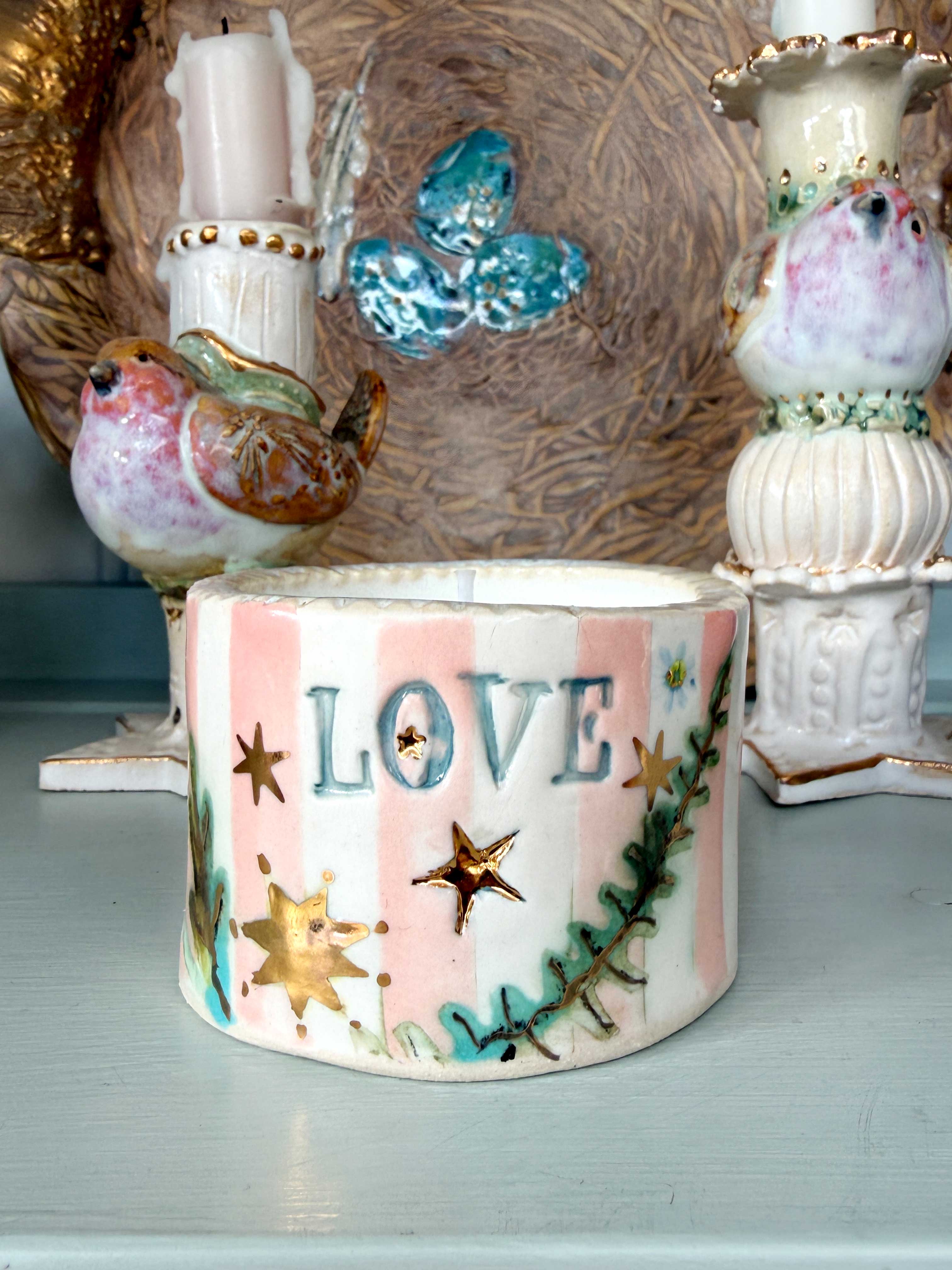 A handmade ceramic container with the word 'LOVE' in raised blue letters, decorated with pink and white stripes, gold stars, and green pine branches, displayed on a shelf with vintage bird candleholders.