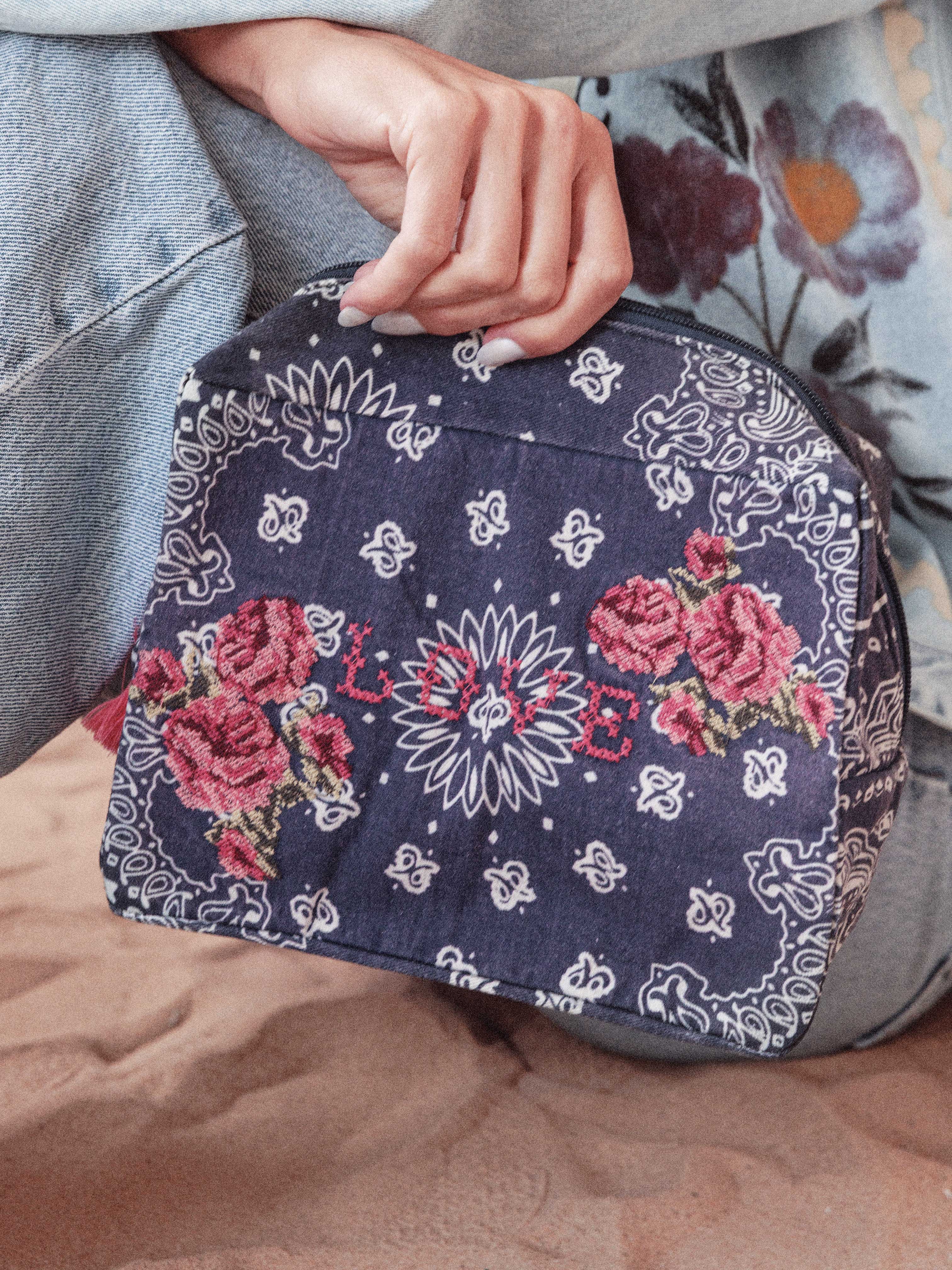 Person holding a navy bandana print canvas cosmetic bag embroidered with cross stitch roses and the word "LOVE."