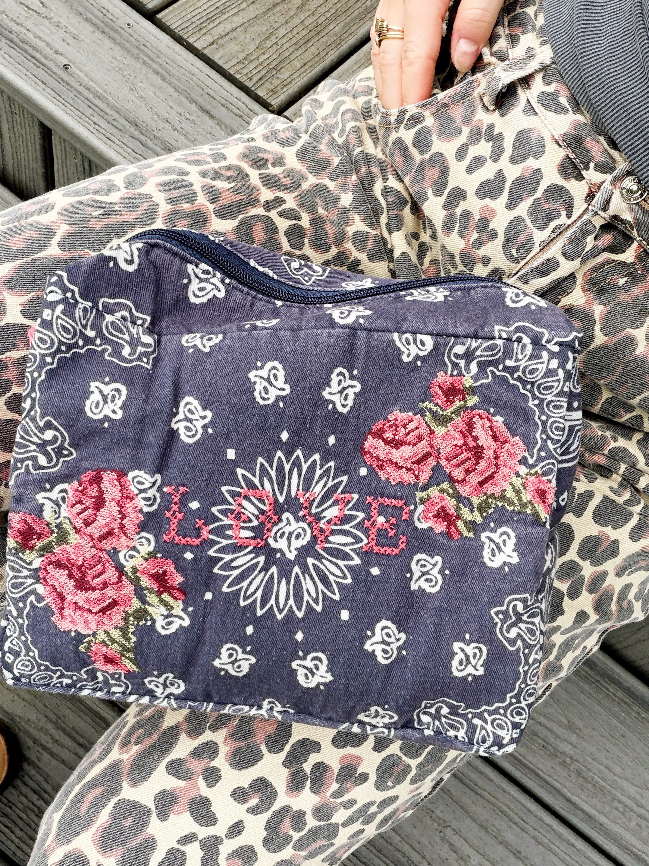 Navy bandana print canvas cosmetic bag embroidered with cross stitch roses and the word "LOVE," styled on a woman's lap with leopard print jeans.
