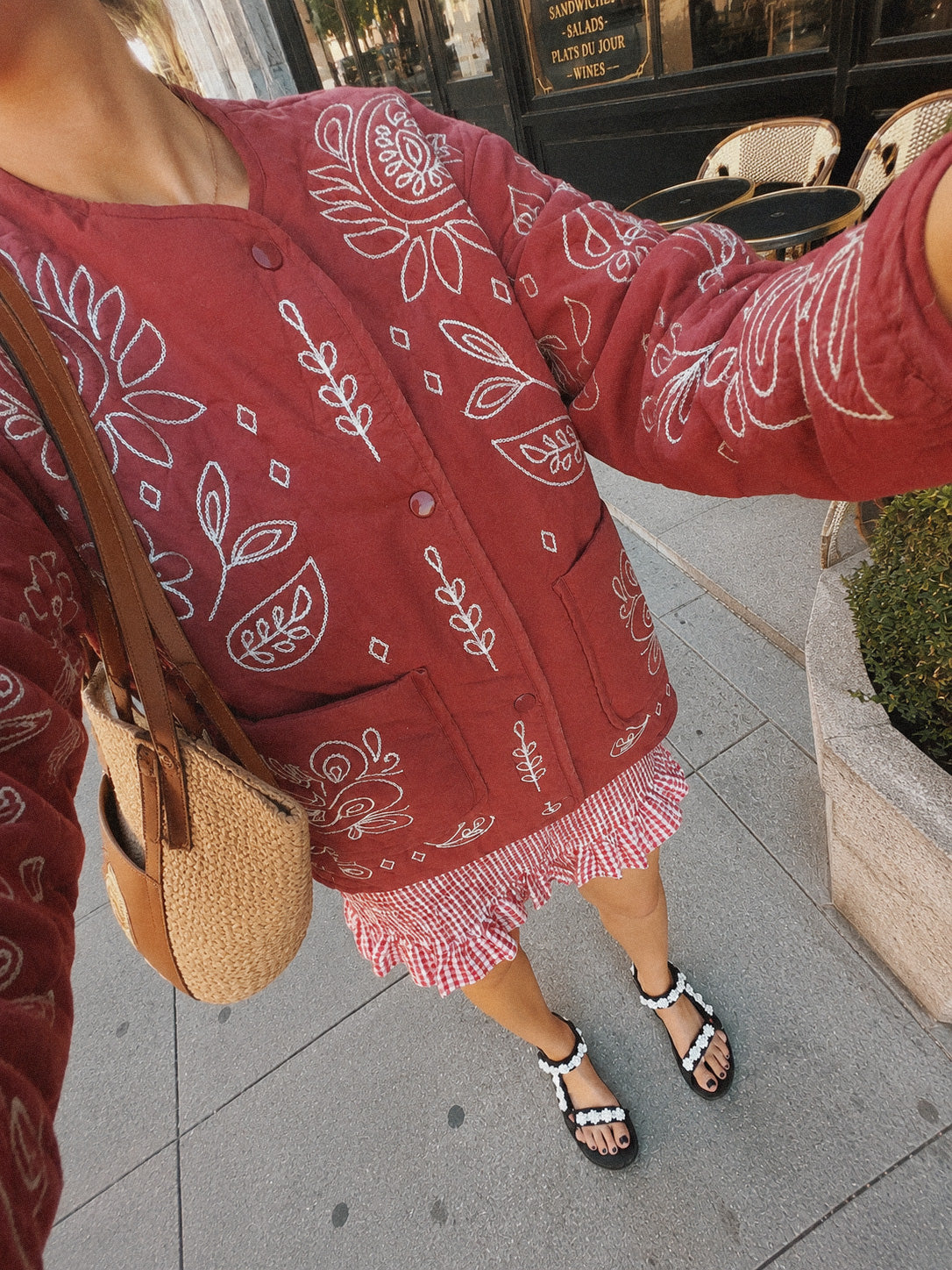 Woman wearing a burgundy quilted jacket with cream bandana-inspired embroidery, styled with a gingham skirt and woven shoulder bag.