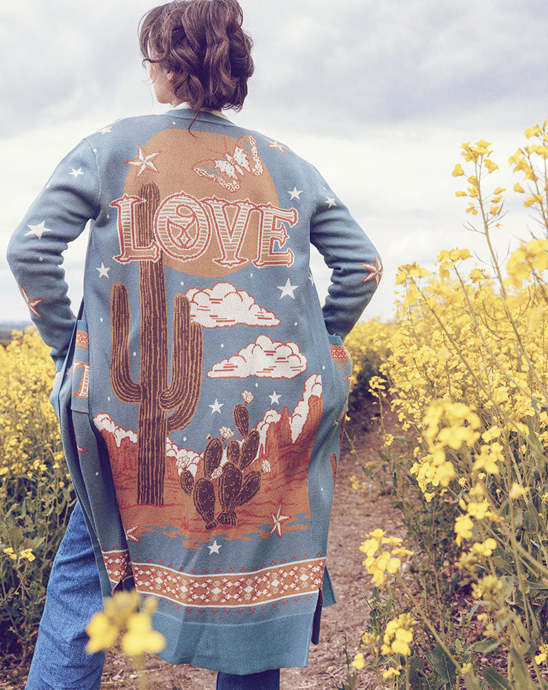 Back view of a person in a field of yellow flowers wearing a teal long knit cardigan featuring a western desert landscape knit design with a large cactus, stars, clouds, mesas, and the word "LOVE" on the back.