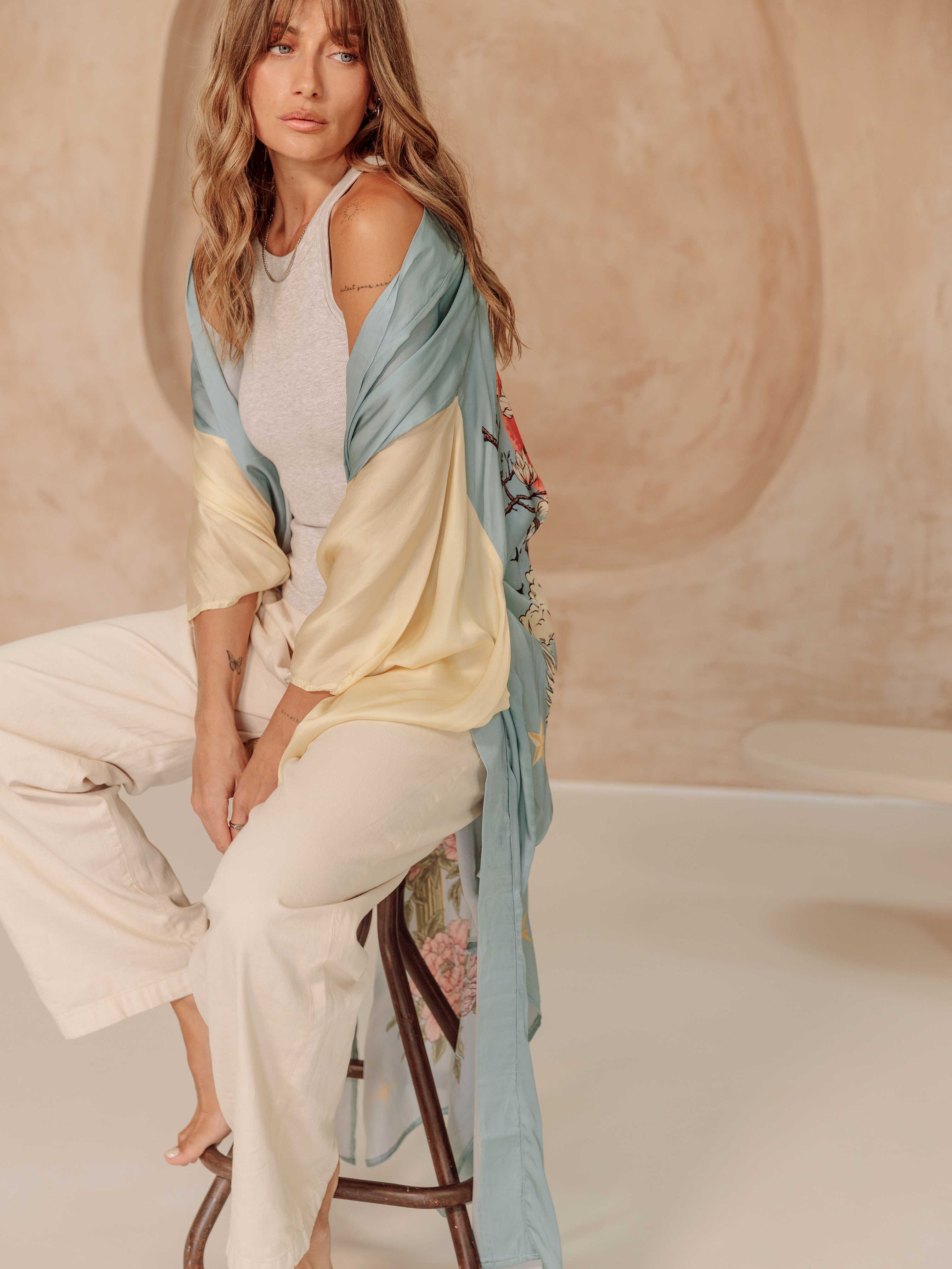 Woman wearing a light blue kimono with cream sleeves and floral print accents, layered over a grey tank top and cream pants.