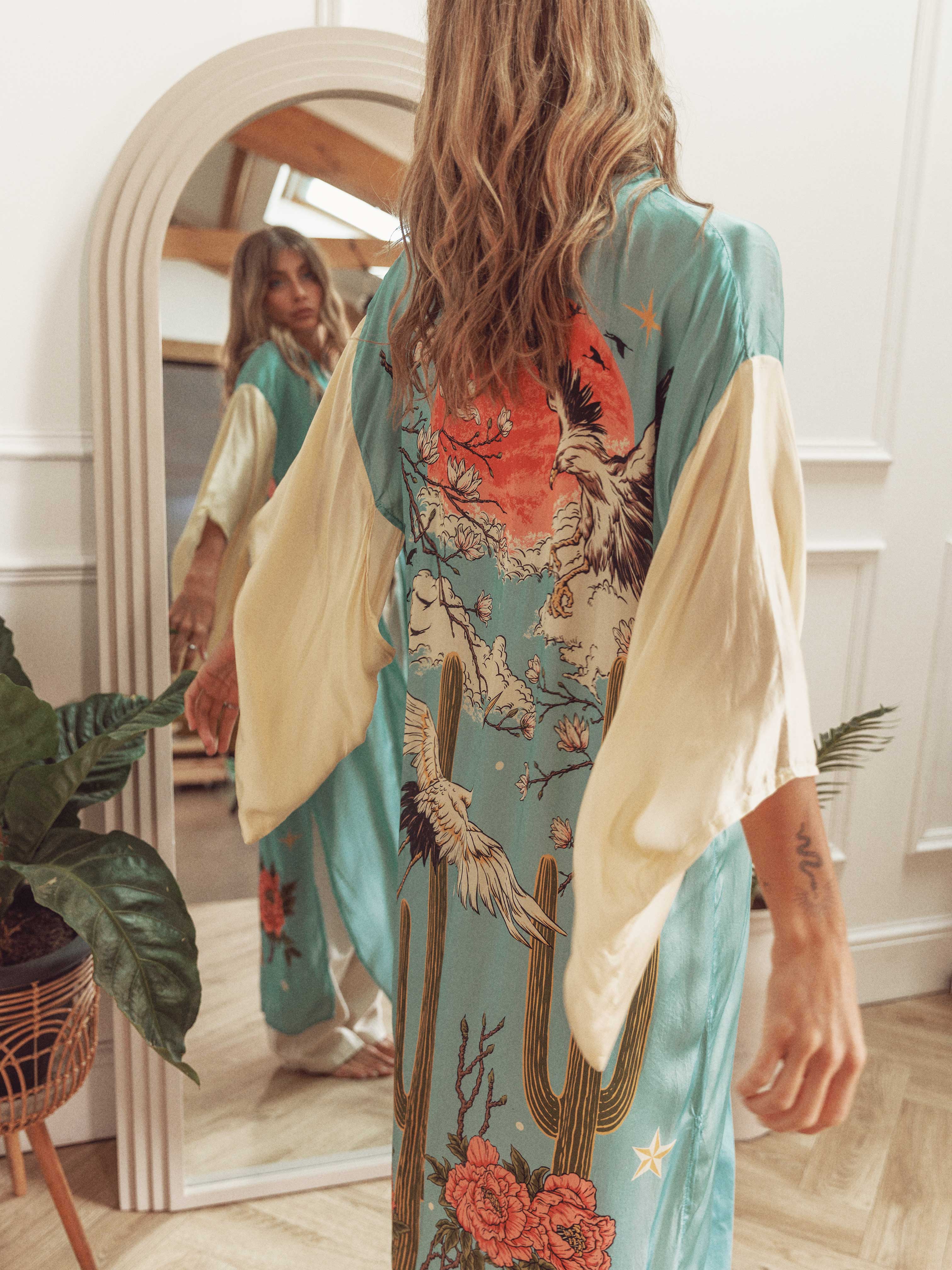 Back view of a light blue kimono with cream sleeves featuring a crane, red sun, cacti, and floral print, reflected in a mirror.