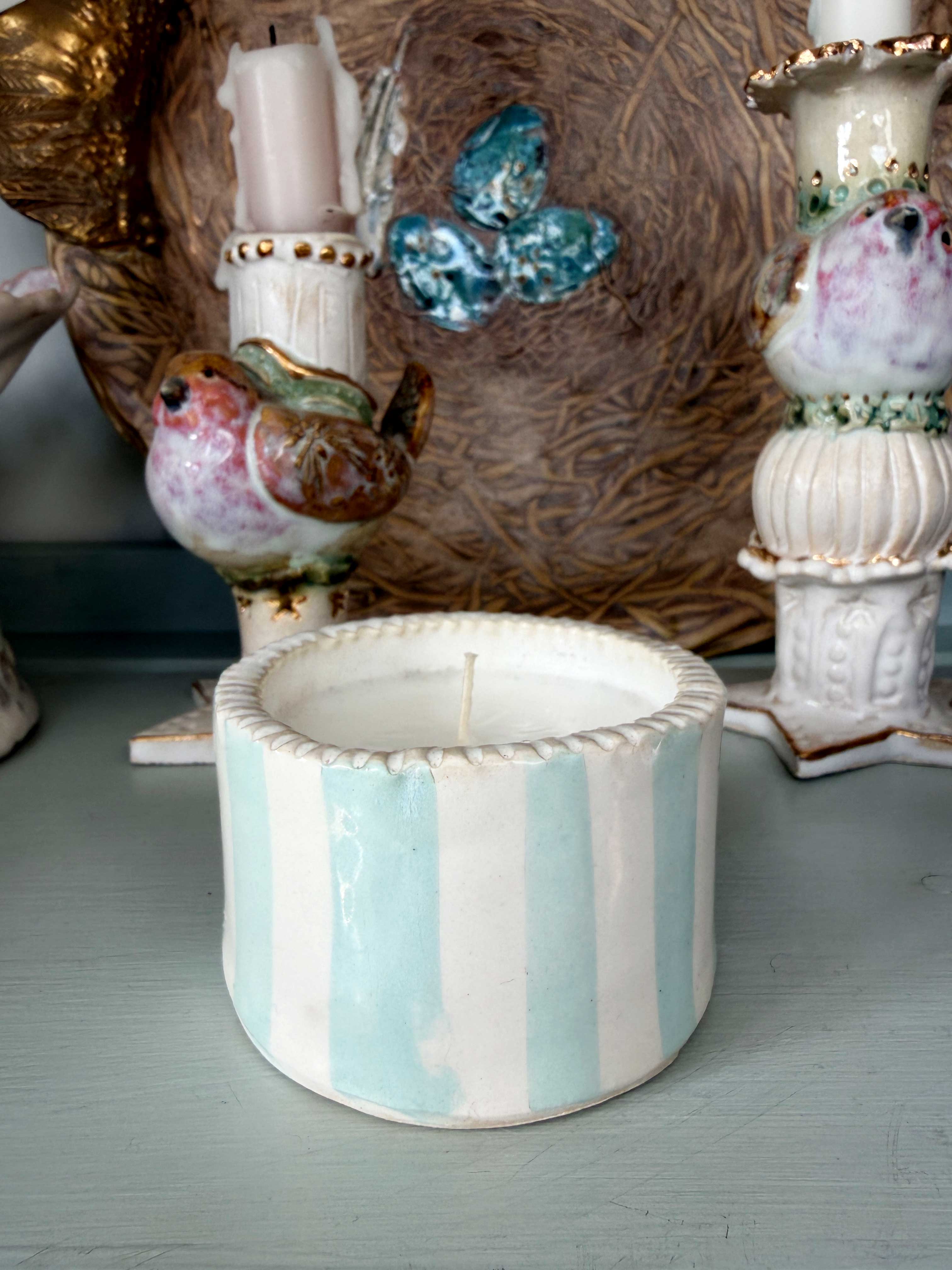 A small cylindrical ceramic candle holder with vertical mint green and white stripes, holding a white candle, displayed on a light blue-gray surface with other decorative ceramic items in the background.