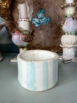 A small cylindrical ceramic candle holder with vertical mint green and white stripes, holding a white candle, displayed on a light blue-gray surface with other decorative ceramic items in the background.