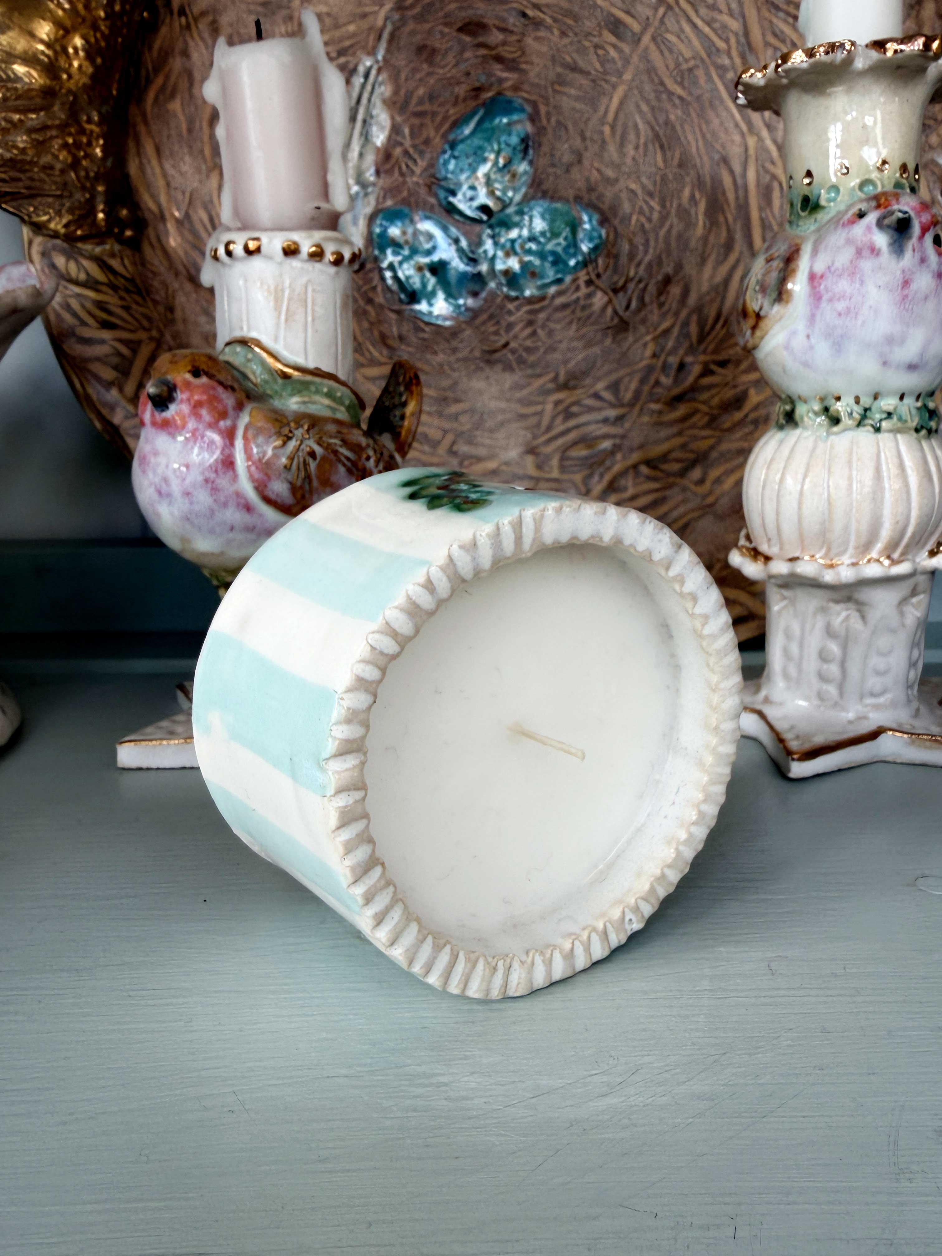 A small ceramic candle holder with a scalloped rim, featuring alternating mint green and white stripes, shown lying on its side on a painted blue-gray surface.